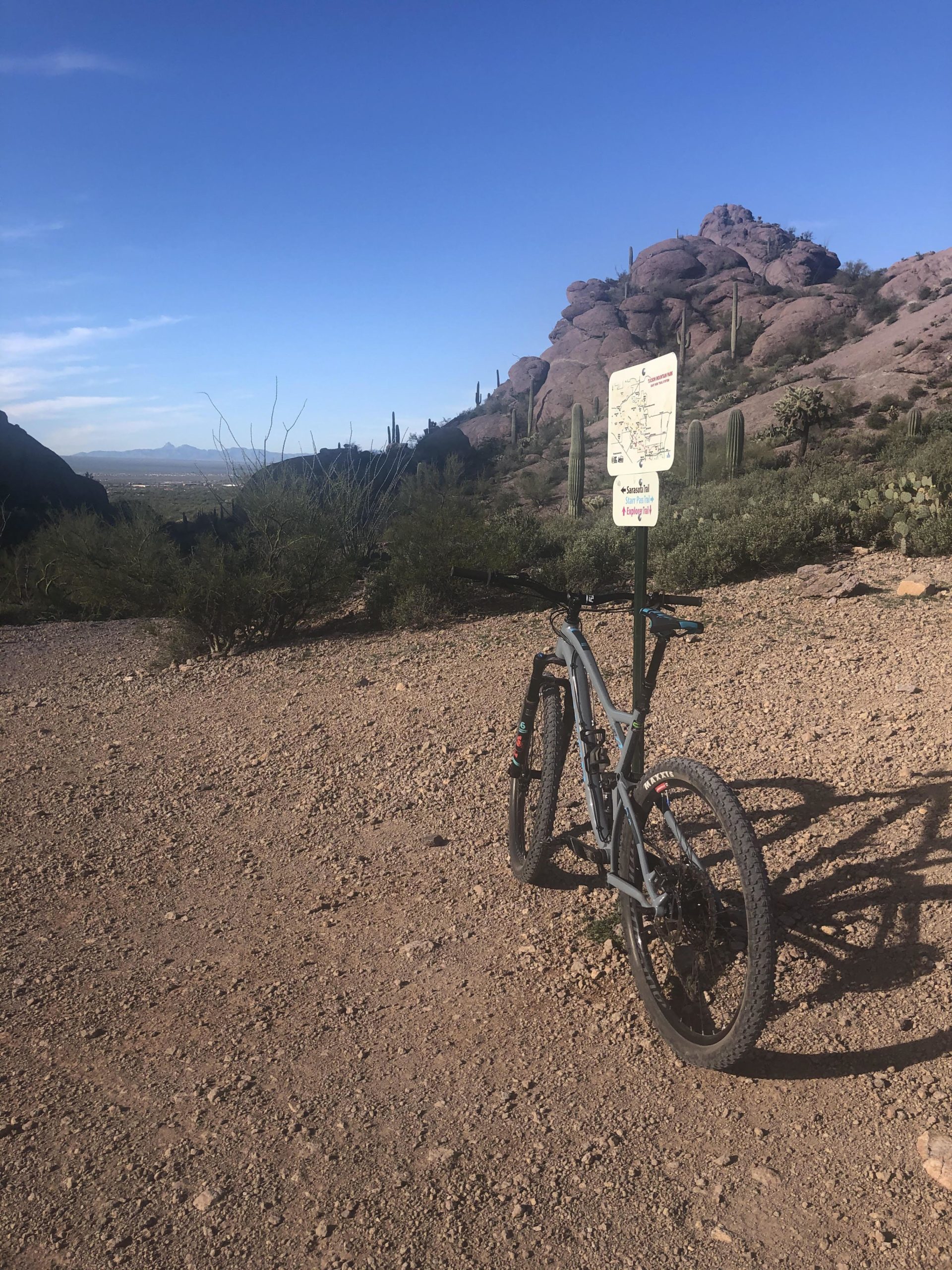 A mountain bike parked on a gravel trail in a desert landscape, with large rock formations and cacti in the background. A trail sign can be seen nearby, providing information about the area. The sky is clear and blue, suggesting a sunny day. Tucson Mountain Park mountain bike trail.