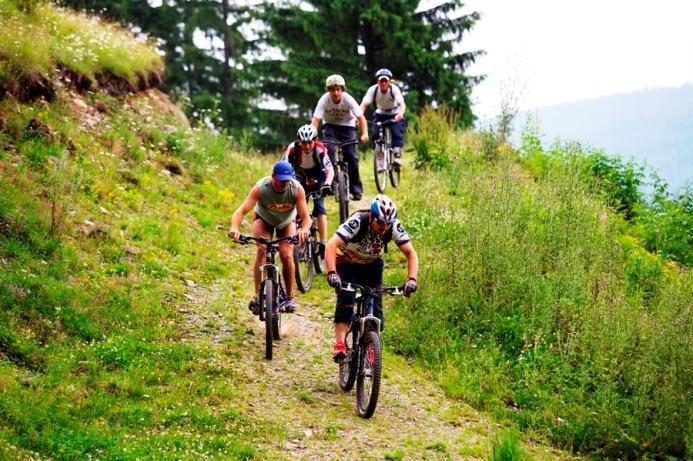 Five mountain bikers riding uphill on a grassy trail surrounded by trees. Some bikers are wearing helmets and various athletic clothing, with one cyclist in the foreground leaning forward as they navigate the incline. The background features a lush green landscape with distant hills.