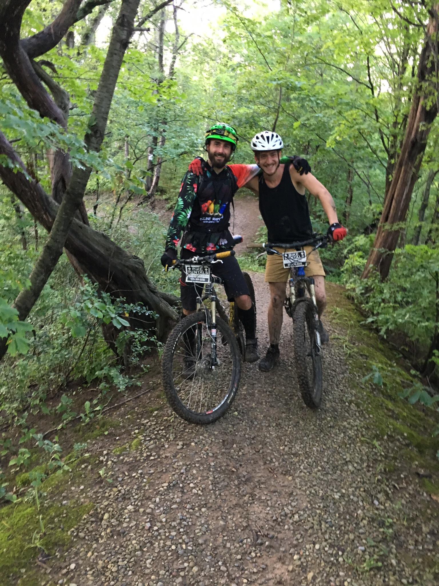 Two mountain bikers pose for a photo on a gravel trail surrounded by green trees. One rider is wearing a colorful long-sleeve jersey and a green helmet, while the other is in a sleeveless black shirt and a white helmet. They are both smiling and leaning on their bikes, showing a friendly camaraderie.