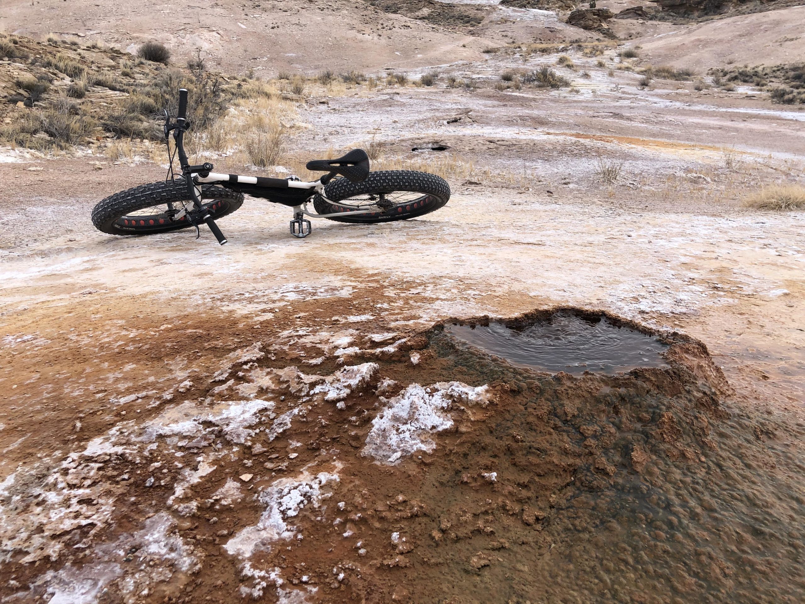 A fat-tire bike lays on its side beside a small, muddy puddle in a dry, rugged landscape. The ground is a mix of reddish-brown dirt and white salt deposits, with sparse vegetation in the background. The scene conveys a remote outdoor environment, likely in a desert or arid area. White Ridge Bike Trails mountain bike trail.