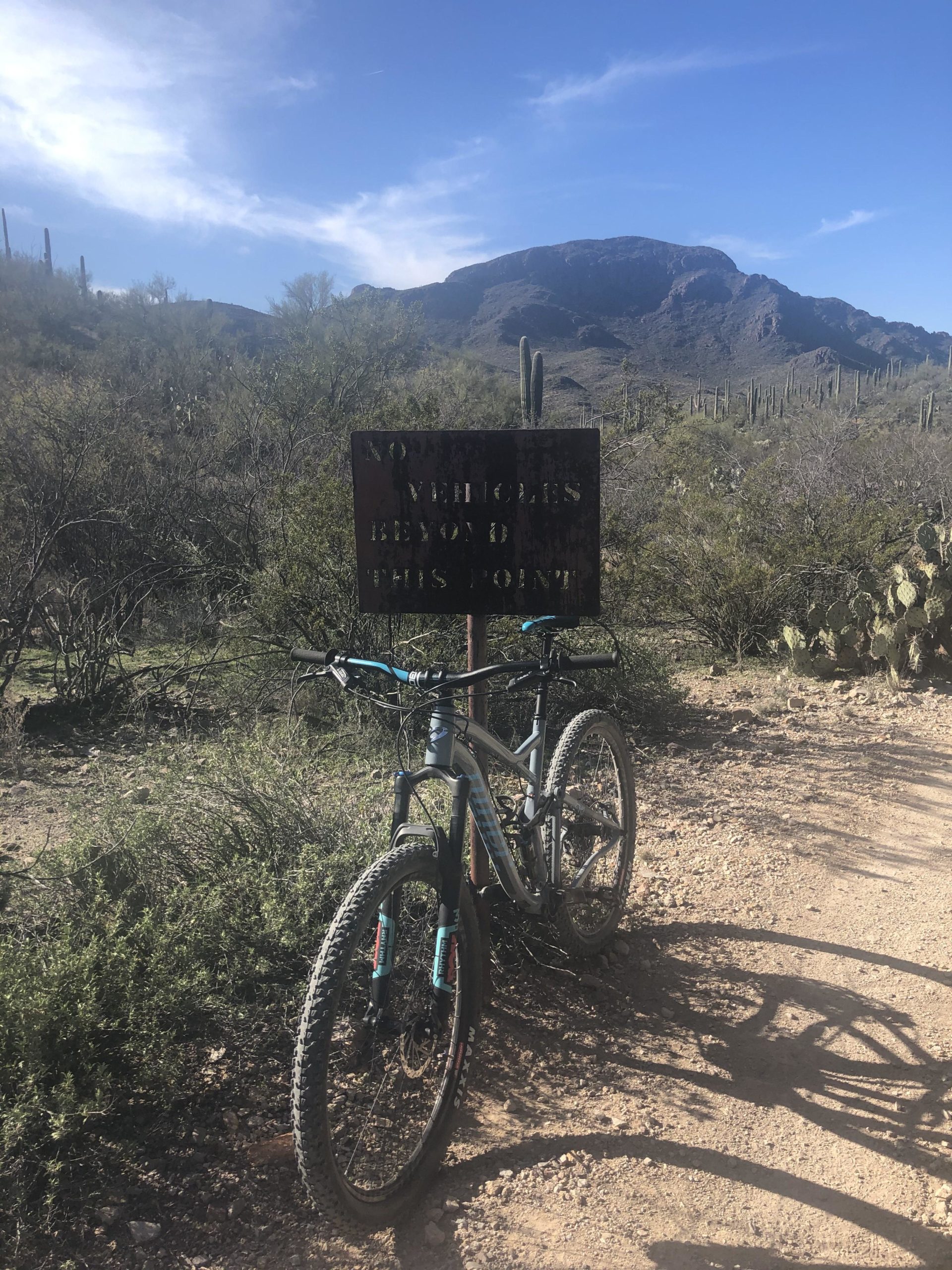 A mountain bike parked next to a weathered sign that reads "No Vehicles Beyond This Point," with a mountainous landscape and cacti in the background under a blue sky. Tucson Mountain Park mountain bike trail.