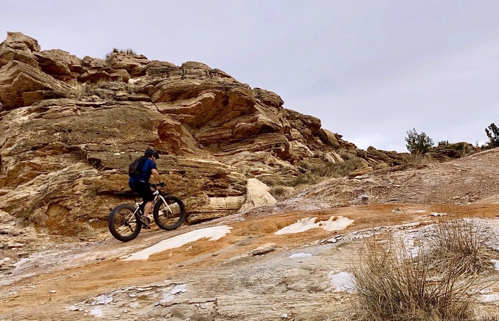 A person riding a fat tire mountain bike on a rocky trail, surrounded by large rock formations and sparse vegetation. The landscape features earthy tones with a cloudy sky above. White Ridge Bike Trails mountain bike trail.