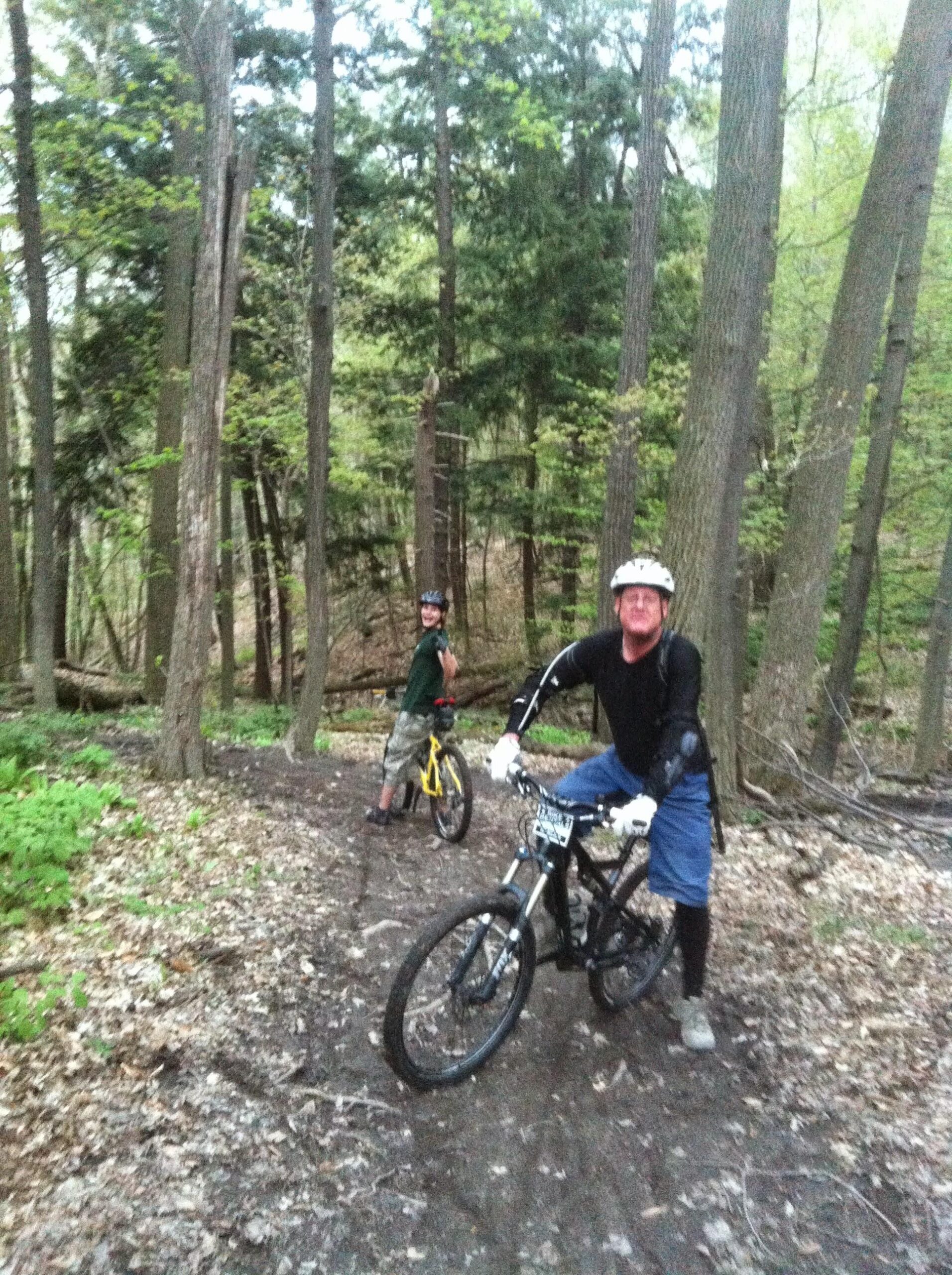 Two people on mountain bikes are positioned on a dirt trail in a forest. One rider, wearing a black shirt and helmet, is standing next to a black mountain bike, while the other, dressed in a green shirt and yellow bike, is looking back with a smile. The scene is surrounded by tall trees and lush greenery, indicating a spring setting.