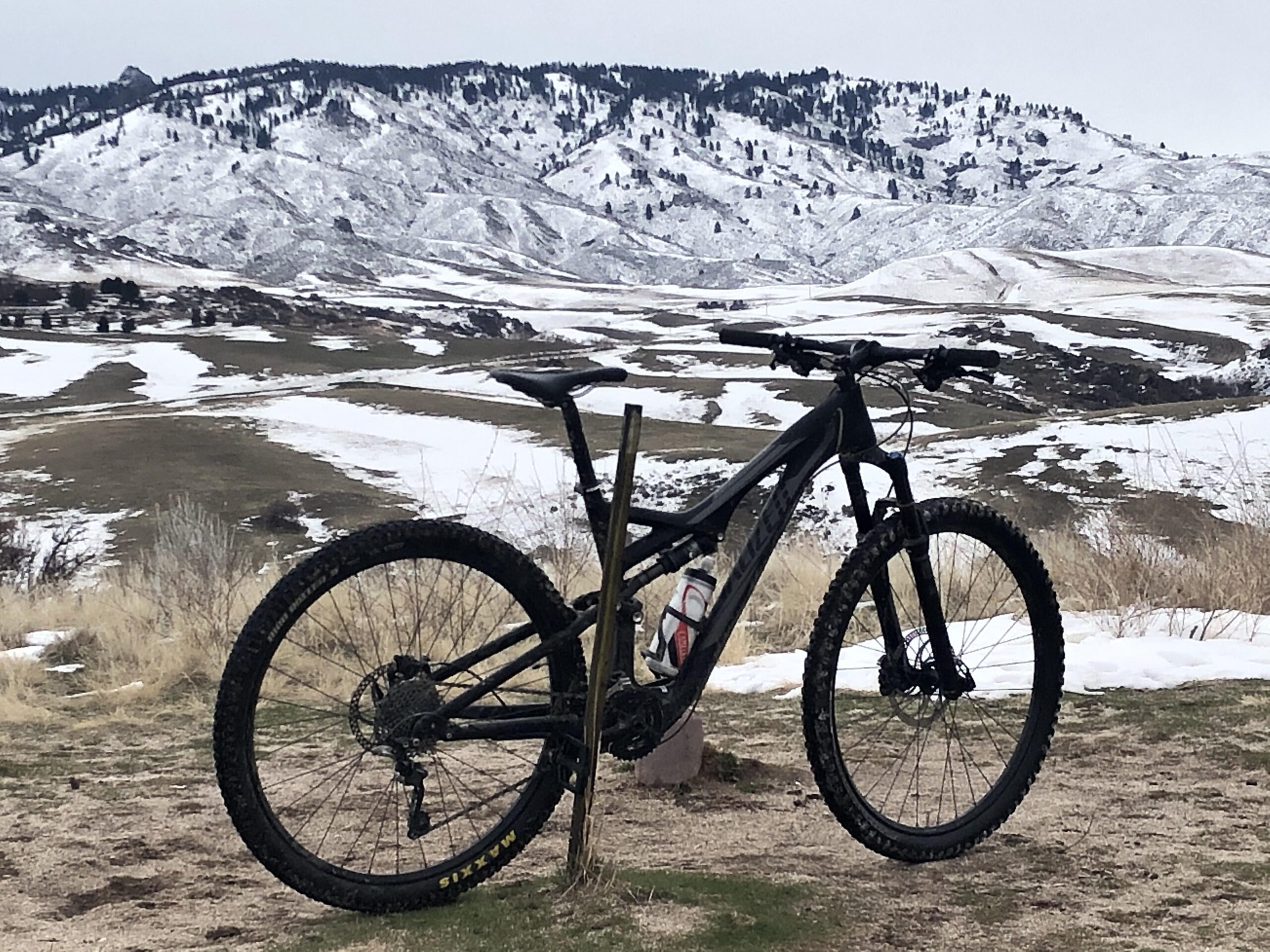 Specialized Stumpjumper FSR: A black mountain bike parked on a dirt path, with snow-covered mountains in the background. The landscape features patches of snow and grassy areas under a cloudy sky.