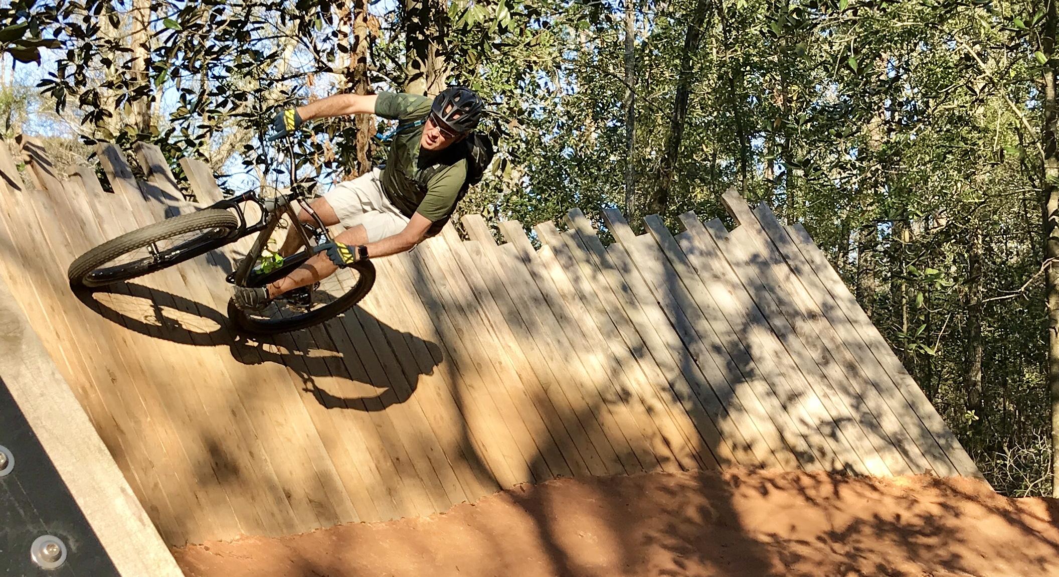 A mountain biker performing a sharp turn on a wooden ramp in a forested area, wearing a helmet and gloves. The rider is positioned at an angle, skillfully balancing on the bike as they navigate the ramp. Sunlight filters through the trees, casting dappled shadows on the ground. Beaver Creek / Forever Wild Trails mountain bike trail.