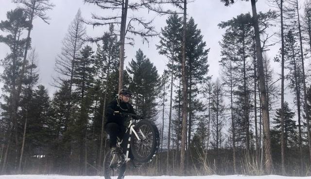 A cyclist performs a wheelie on a fat bike in a snowy forest, surrounded by tall, leafless trees under a cloudy sky. Spencer Mountain mountain bike trail.