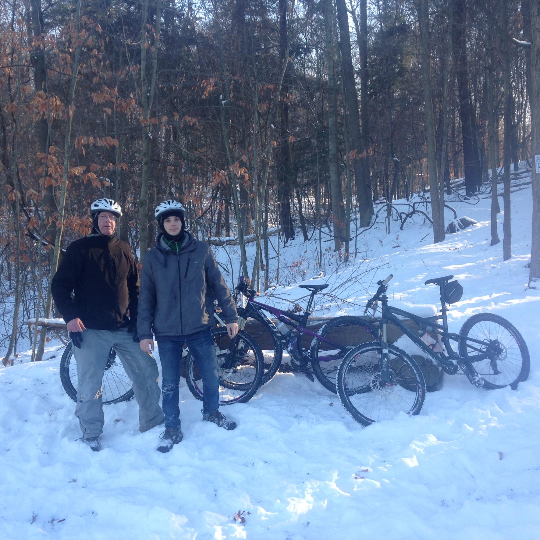 Two individuals standing in a snowy forest, both wearing helmets and winter clothing. They are positioned beside two mountain bikes resting on the snow. The background features trees with sparse leaves, indicating a winter setting.