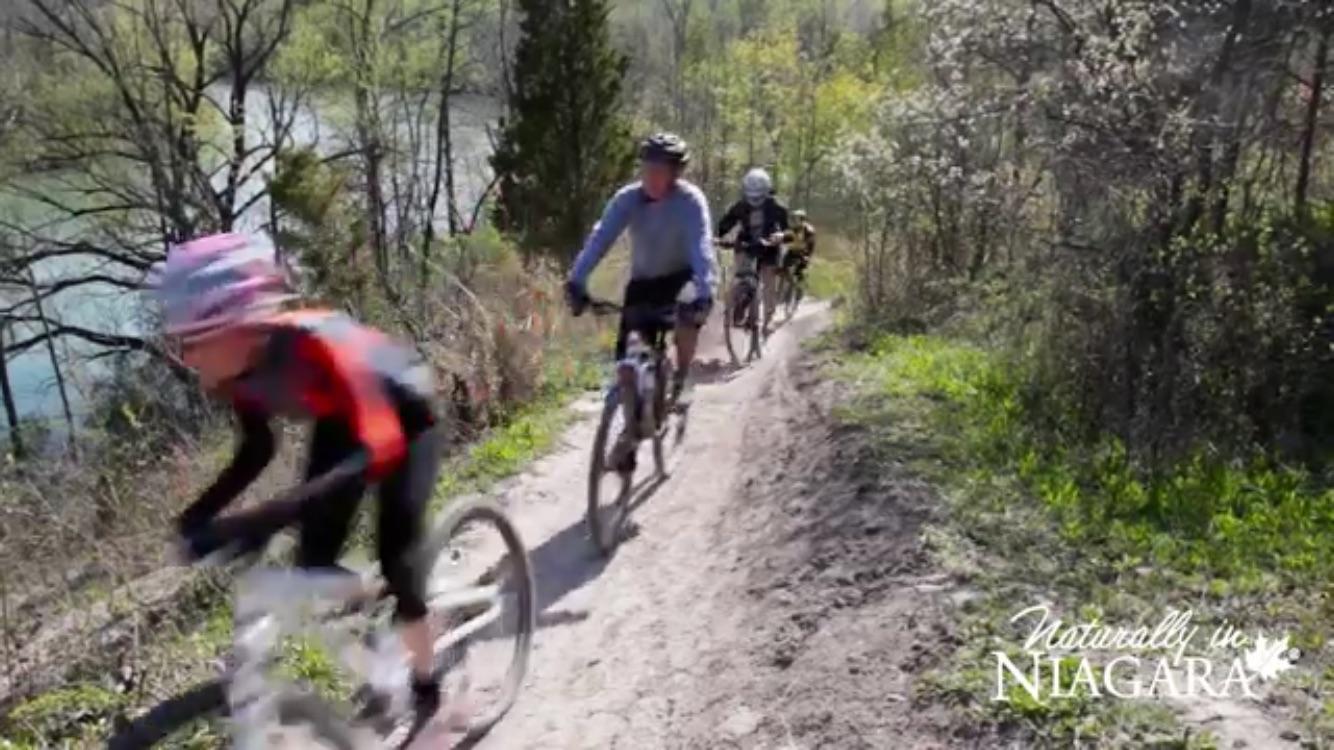 A group of three mountain bikers riding on a dirt trail surrounded by greenery and trees, with a body of water visible in the background on a sunny day.