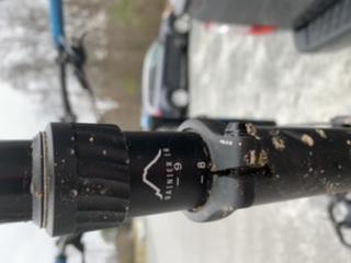 Close-up of a bicycle suspension fork with mud and dirt on it, showing the brand name "Dare" and adjustment markings, set against a blurred background of parked cars and a natural landscape. Blankets Creek mountain bike trail.