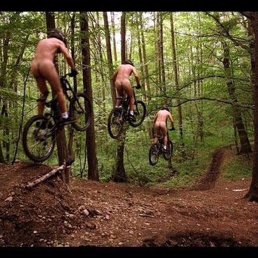 Three cyclists performing jumps on mountain bikes in a forested area. The riders are nude and are captured mid-air as they navigate a dirt trail surrounded by trees.
