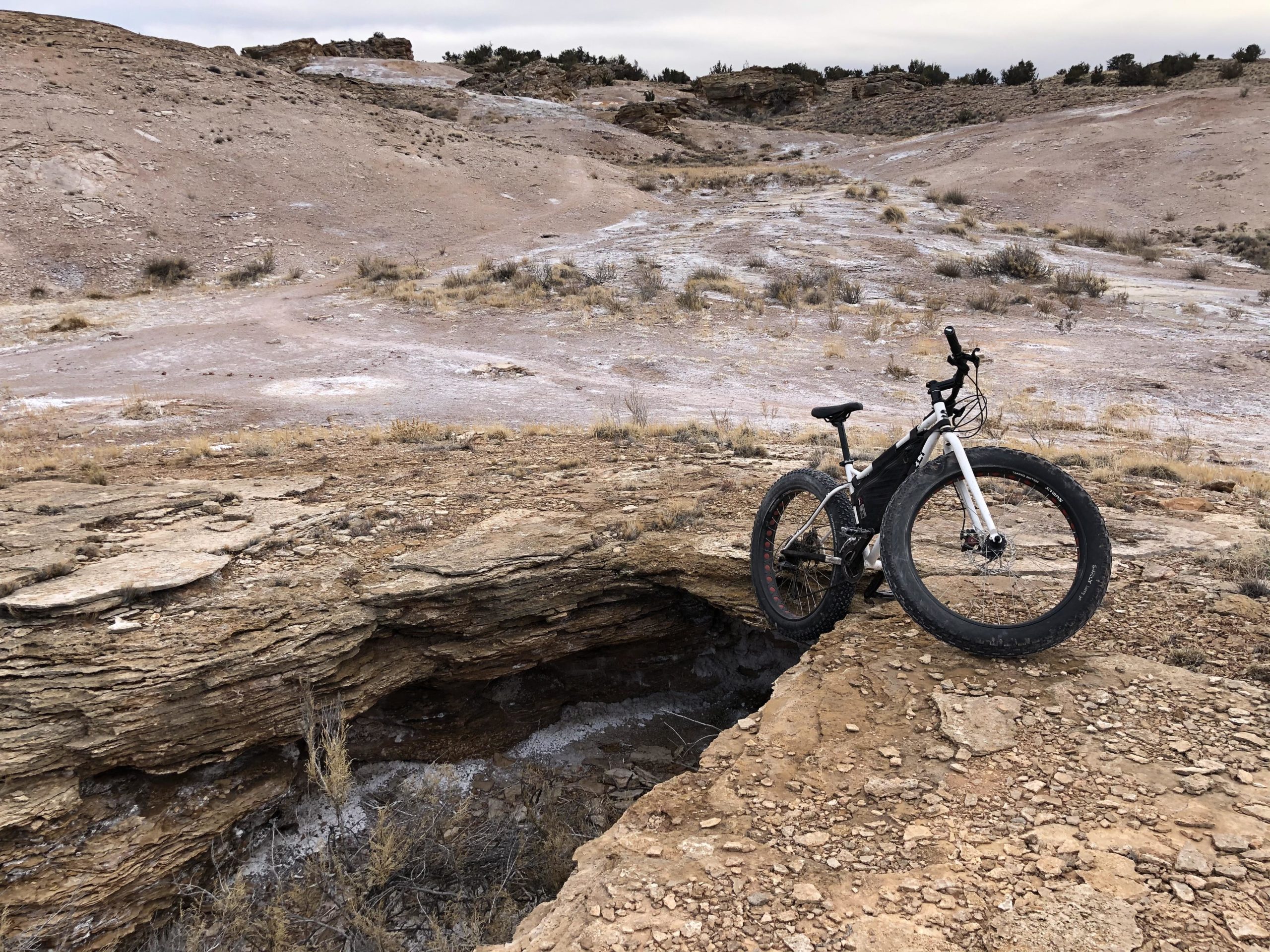 A fat tire bike is parked on the edge of a rocky outcrop overlooking a dry, barren landscape, with patches of sparse vegetation and rugged terrain. The sky is overcast, adding to the desolate atmosphere of the scene. White Ridge Bike Trails mountain bike trail.