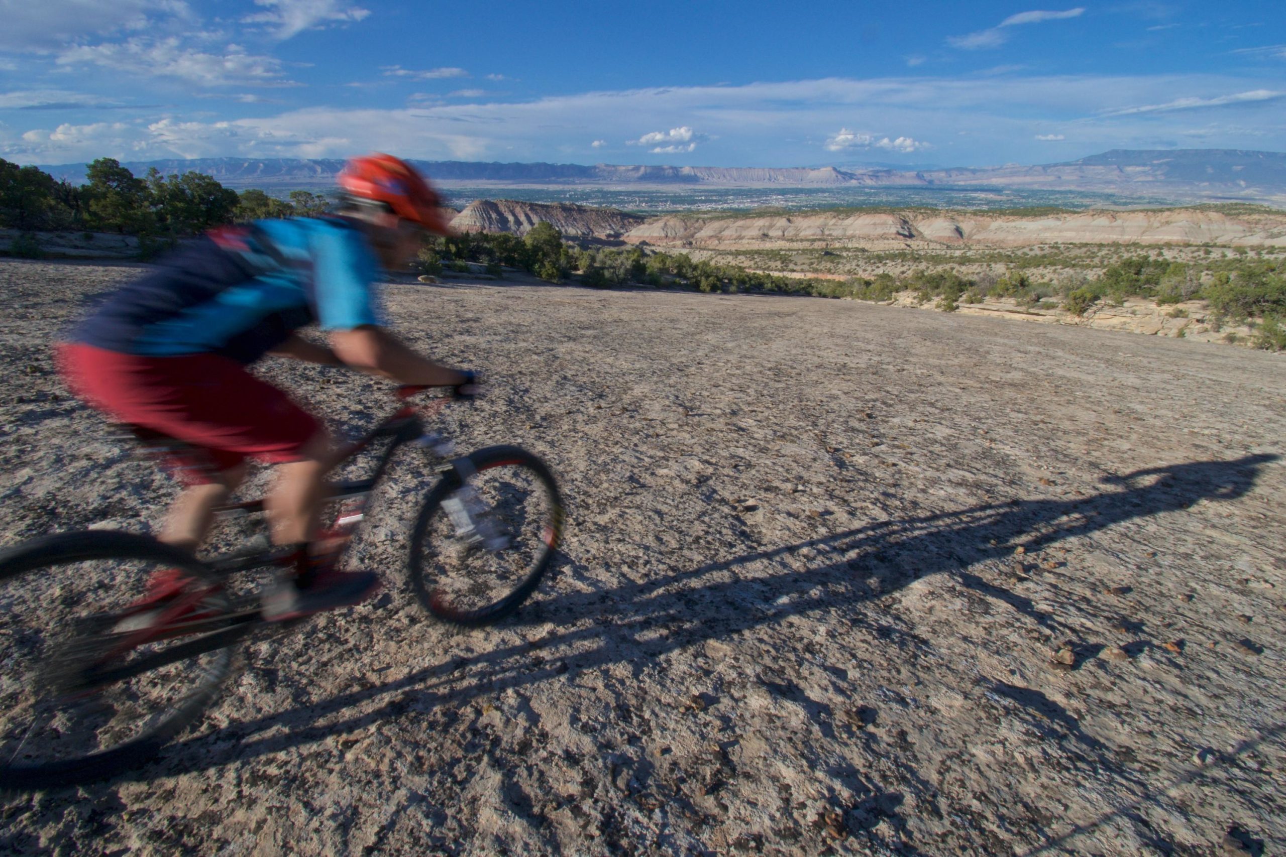 A blurred image of a mountain biker riding on a rocky terrain, with a vast landscape of hills and blue skies in the background. The biker is wearing a red helmet and a blue and black jersey, creating a sense of motion as they speed across the rugged surface. The Ribbon mountain bike trail.