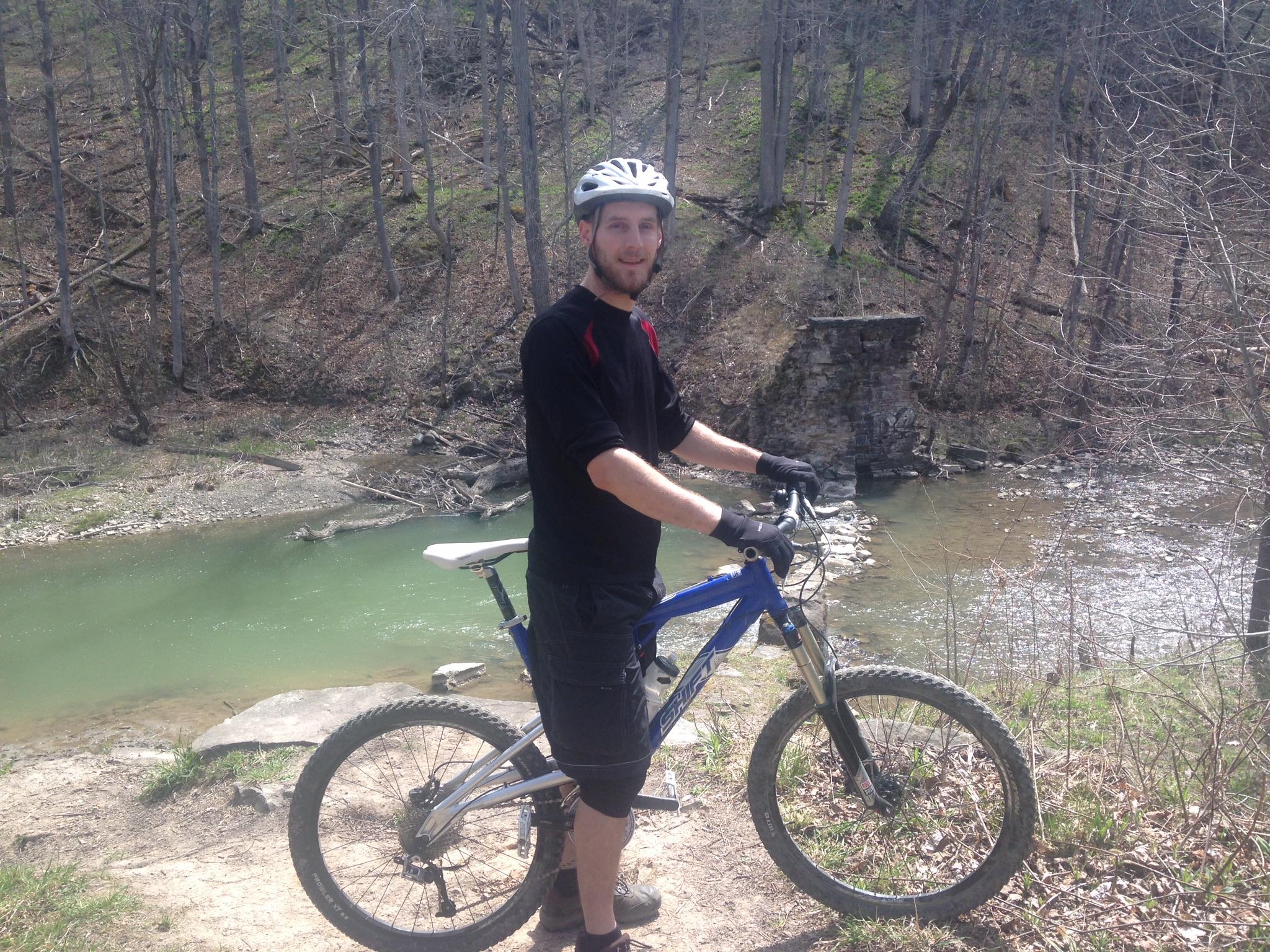 A person in a black shirt and helmet stands next to a blue mountain bike on a dirt path beside a river, with trees and rocky terrain in the background. The scene is set in a natural outdoor environment during springtime.