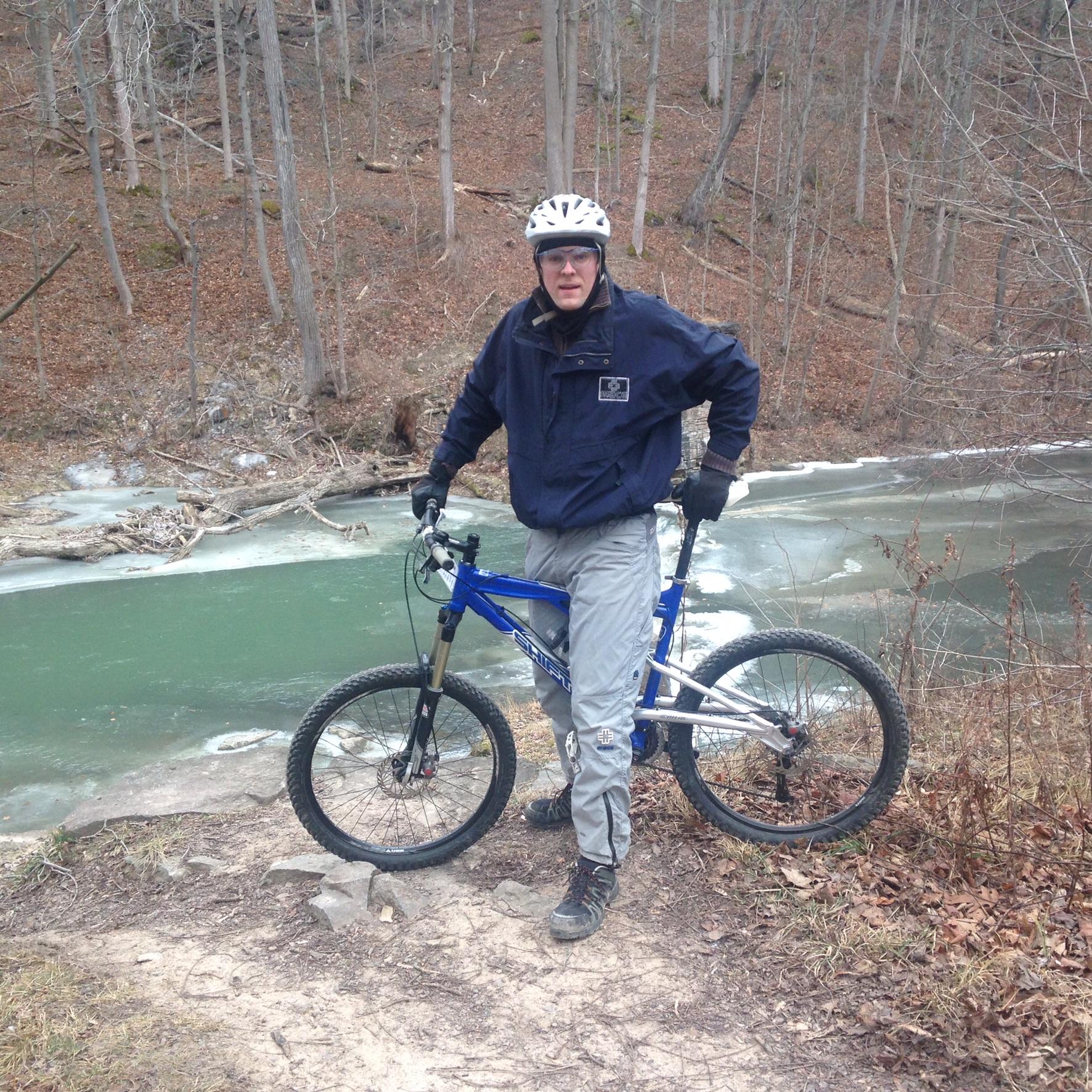 A person wearing a helmet and a dark jacket stands beside a blue mountain bike on a rocky path near a river. The river, partially frozen, is surrounded by trees with brown leaves scattered on the ground, indicating a late autumn or early winter setting.