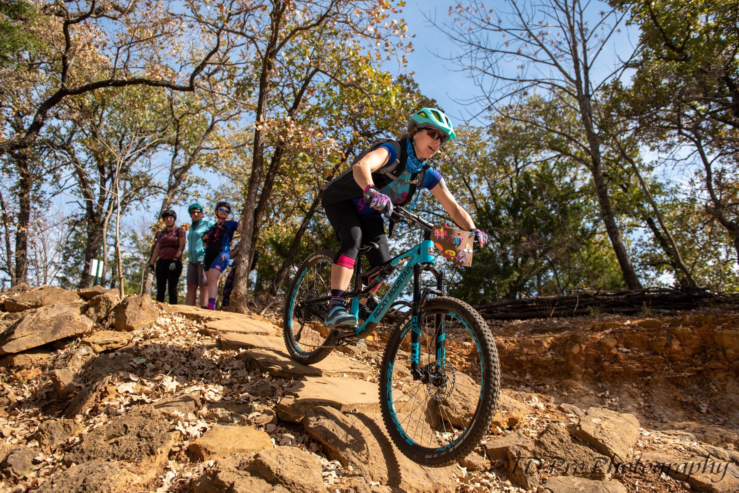 A young girl skillfully rides a mountain bike down a rocky terrain, surrounded by trees with autumn foliage. In the background, three friends, also wearing helmets, observe her ride. The scene captures the excitement of biking and the camaraderie of outdoor activities.