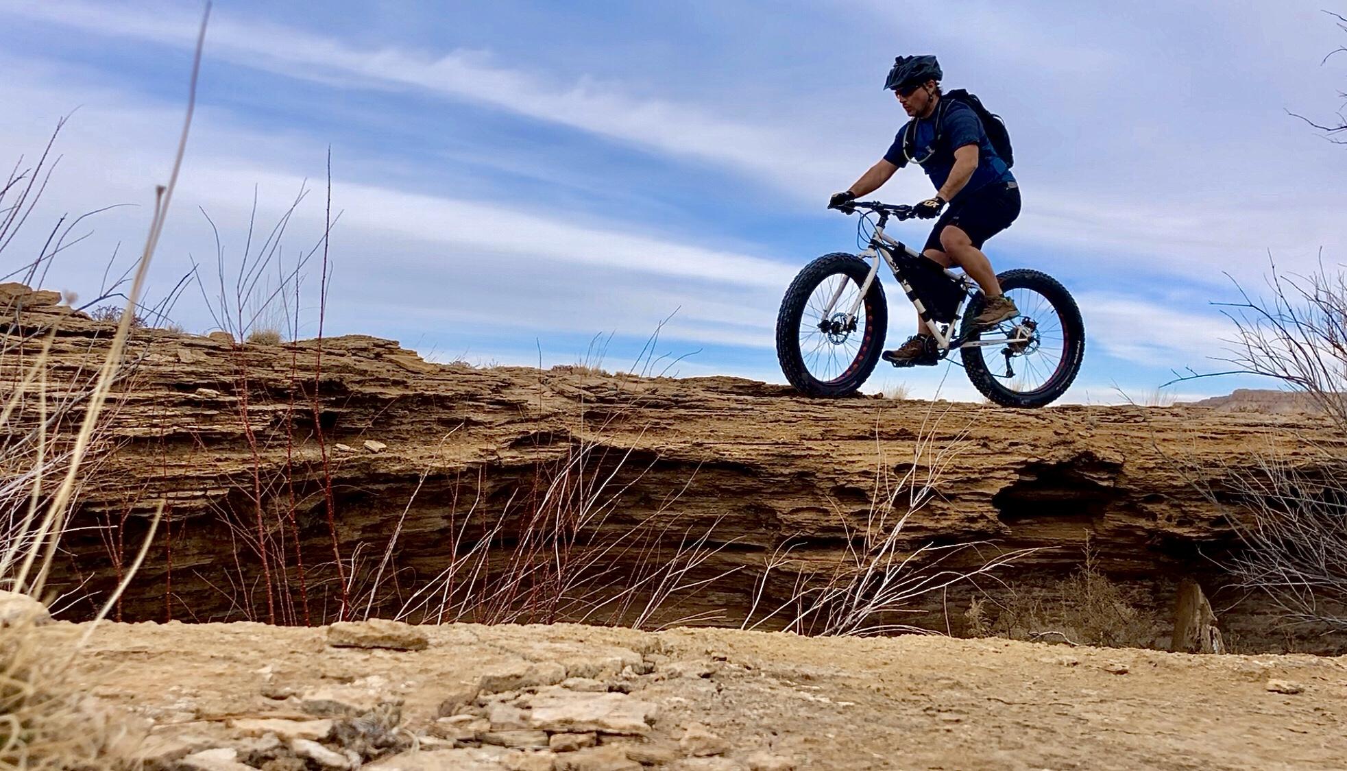 A cyclist riding a fat bike on a rocky trail elevated above the ground, with sparse vegetation and a blue sky in the background. The terrain features rugged rock formations and dry grass. White Ridge Bike Trails mountain bike trail.