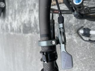 Close-up image of a bicycle handlebar featuring a gear shifter and cable attachment. The gear shifter is mounted on the left side of the handlebar, showing its lever and adjustment mechanism. The background is slightly blurred, focusing on the handlebar components. Blankets Creek mountain bike trail.