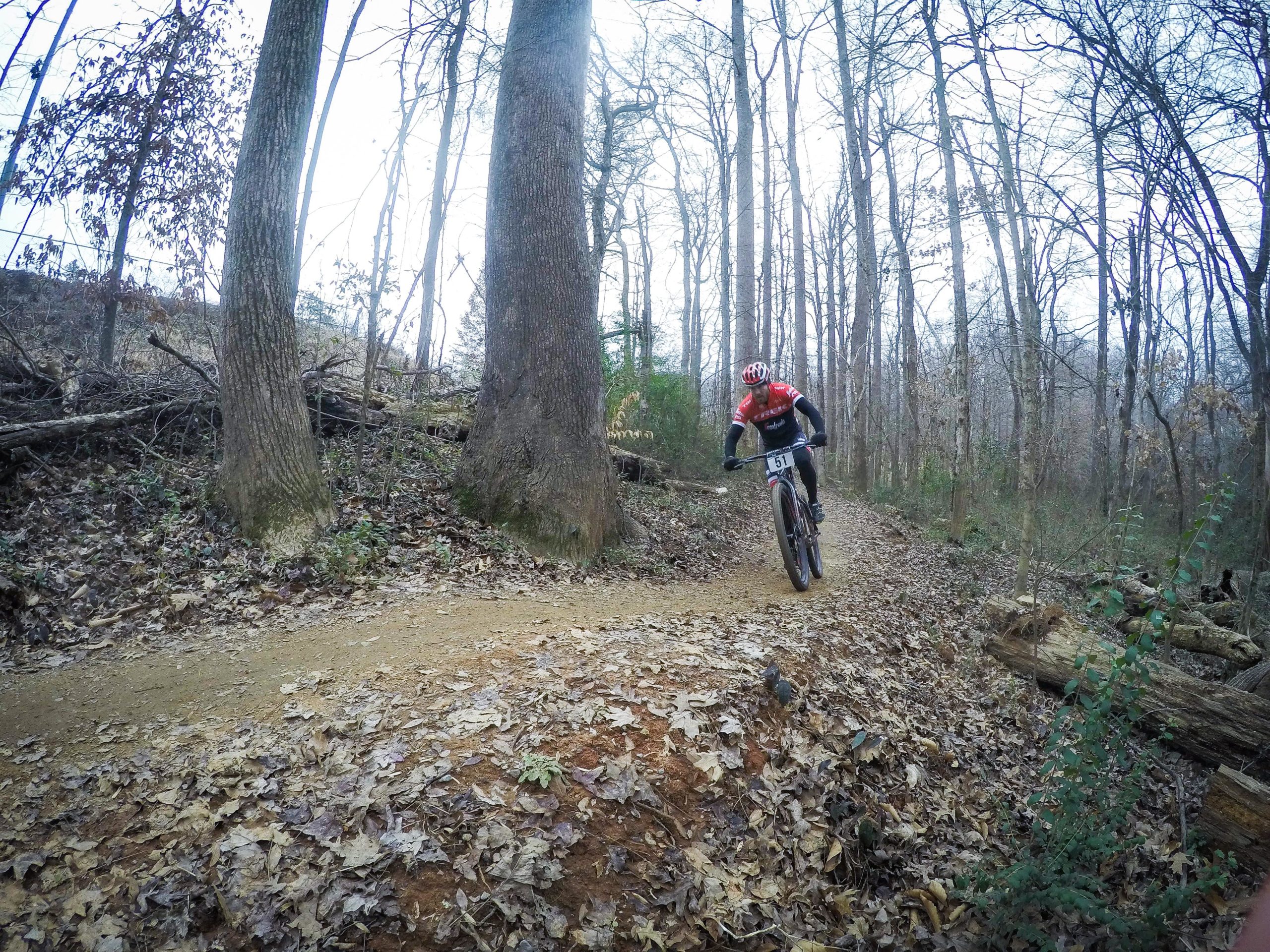 A mountain biker in a red jersey and black pants navigates a winding dirt trail surrounded by tall trees and fallen leaves in a wooded area. The biker is captured mid-action, leaning forward as they ride along the path, with a sense of speed and agility. Finch Park mountain bike trail.