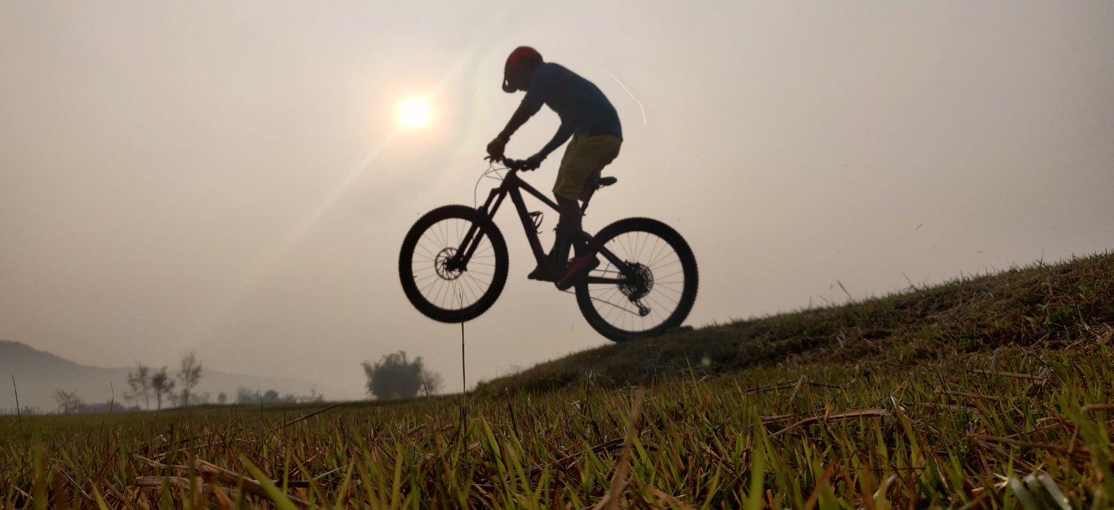 Trek Trek Remedy 8 27.5: A silhouette of a person riding a mountain bike and performing a jump against a hazy background with a setting sun. The foreground features tall grass and a gentle slope leading up to the jump.