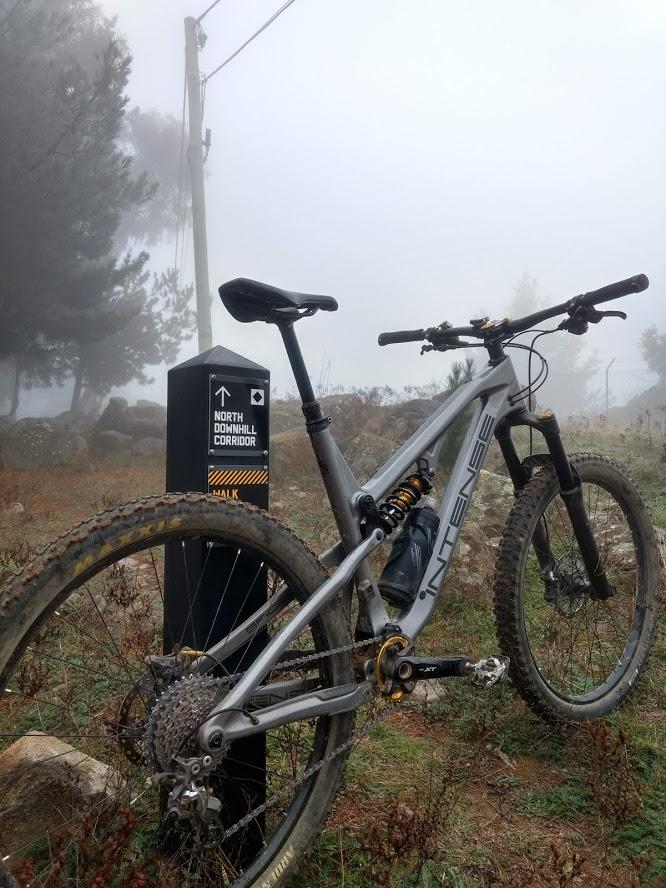 A mountain bike resting on a grassy trail in a foggy environment, with a directional sign nearby indicating the "North Downhill Corridor." The bicycle features a silver frame and is partially covered in dirt, suggesting recent use. The background includes trees and rocky terrain shrouded in mist. Isaacs Ridge mountain bike trail.