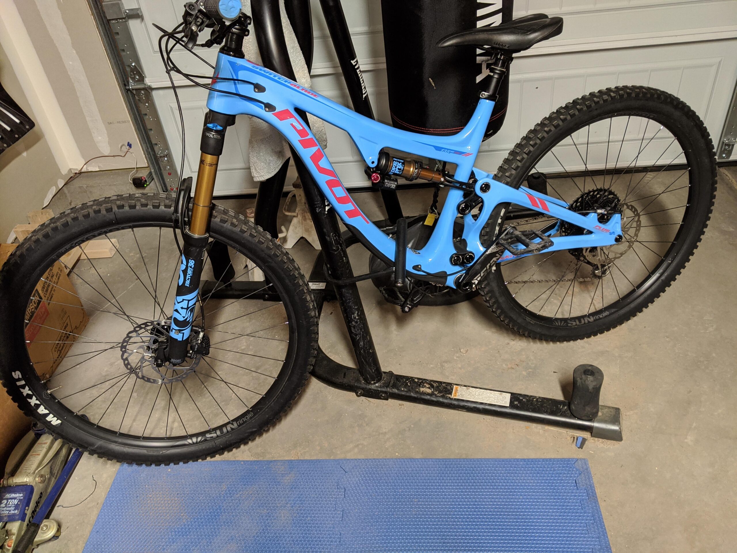 Pivot SwitchBlade: A blue mountain bike with thick tires and a sturdy frame is positioned on a bike repair stand in a garage. The bike features front suspension forks and is equipped with disc brakes. In the background, there are boxes and a heavy bag. The floor is covered with a blue mat.
