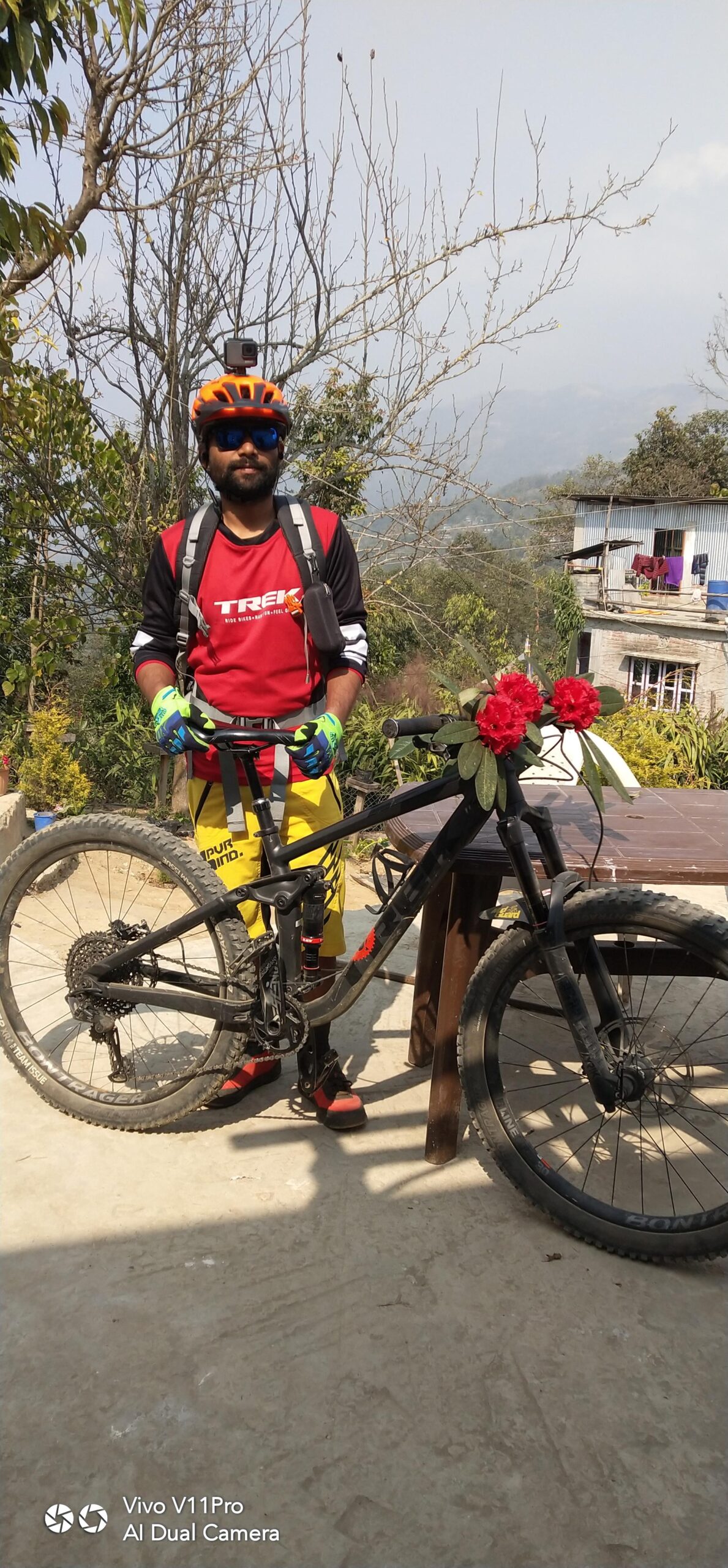 Trek Trek Remedy 8 27.5: A cyclist wearing an orange helmet, sunglasses, and colorful gloves stands next to a mountain bike adorned with flowers. He is dressed in a red and black jersey and yellow shorts, posing on a sunny day with a scenic background of trees and a distant hillside.