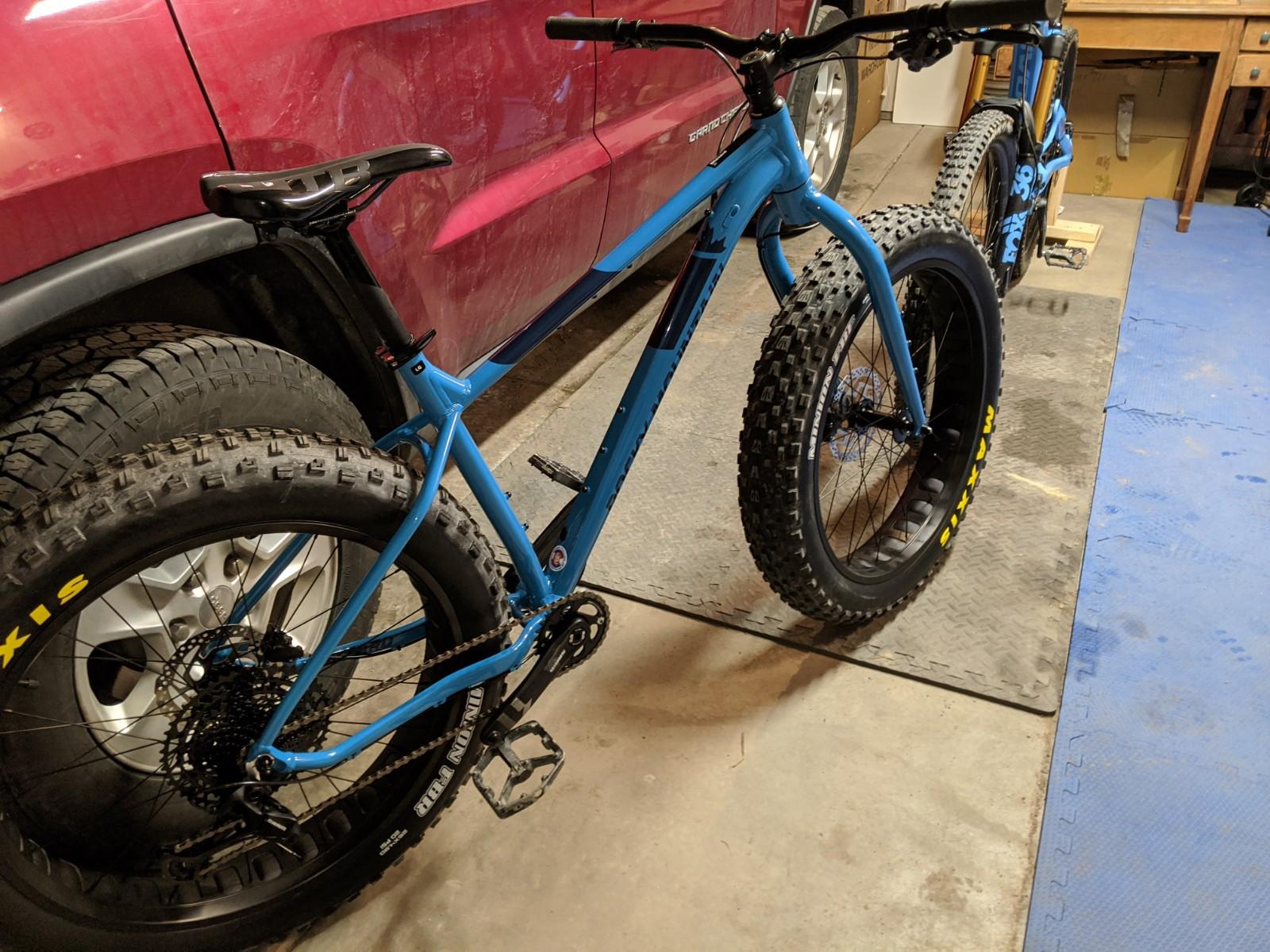 Rocky Mountain Blizzard -20: A blue fat tire bike is parked in a garage next to a red vehicle, with a second bike partially visible in the background. The bike features thick tires and a single front fork, highlighting its design for off-road use. The floor is covered with rubber mats, and a wooden table is seen in the background.