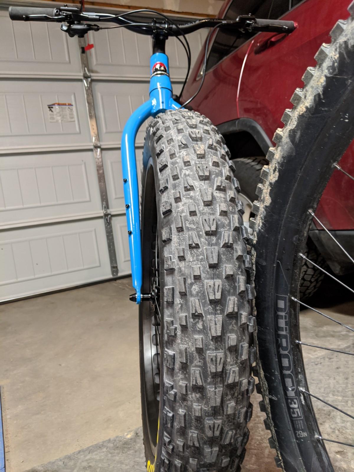 Rocky Mountain Blizzard -20: Close-up view of a blue mountain bike with a thick tire, showing detailed tread patterns, parked in a garage. A red vehicle is partially visible in the background, along with a garage door.