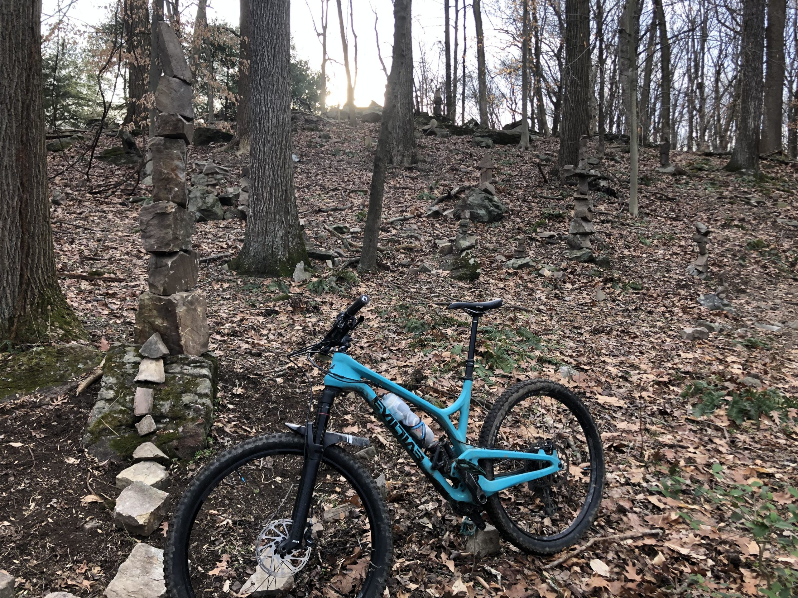 A mountain bike resting on a forest floor covered with leaves, next to a series of stacked stones. The scene is set in a wooded area with tall trees in the background and a faint light peeking through the branches, suggesting early evening. Loch Raven Reservoir mountain bike trail.