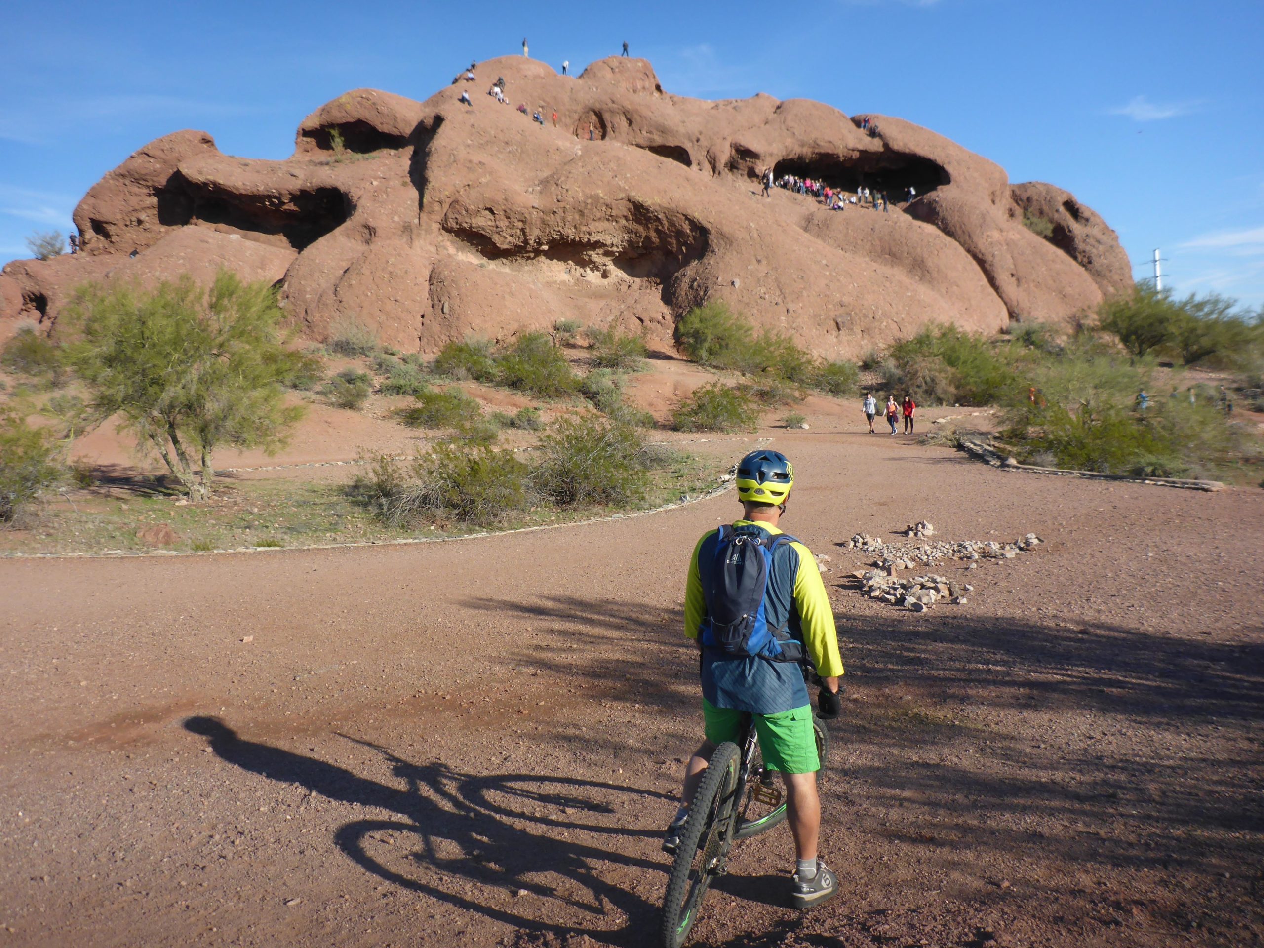 A mountain biker pauses on a dirt path, looking towards a large rock formation with people climbing on its surface. The surrounding landscape features desert vegetation and a blue sky. The scene captures the spirit of outdoor adventure in a scenic environment. Papago Park Area mountain bike trail.