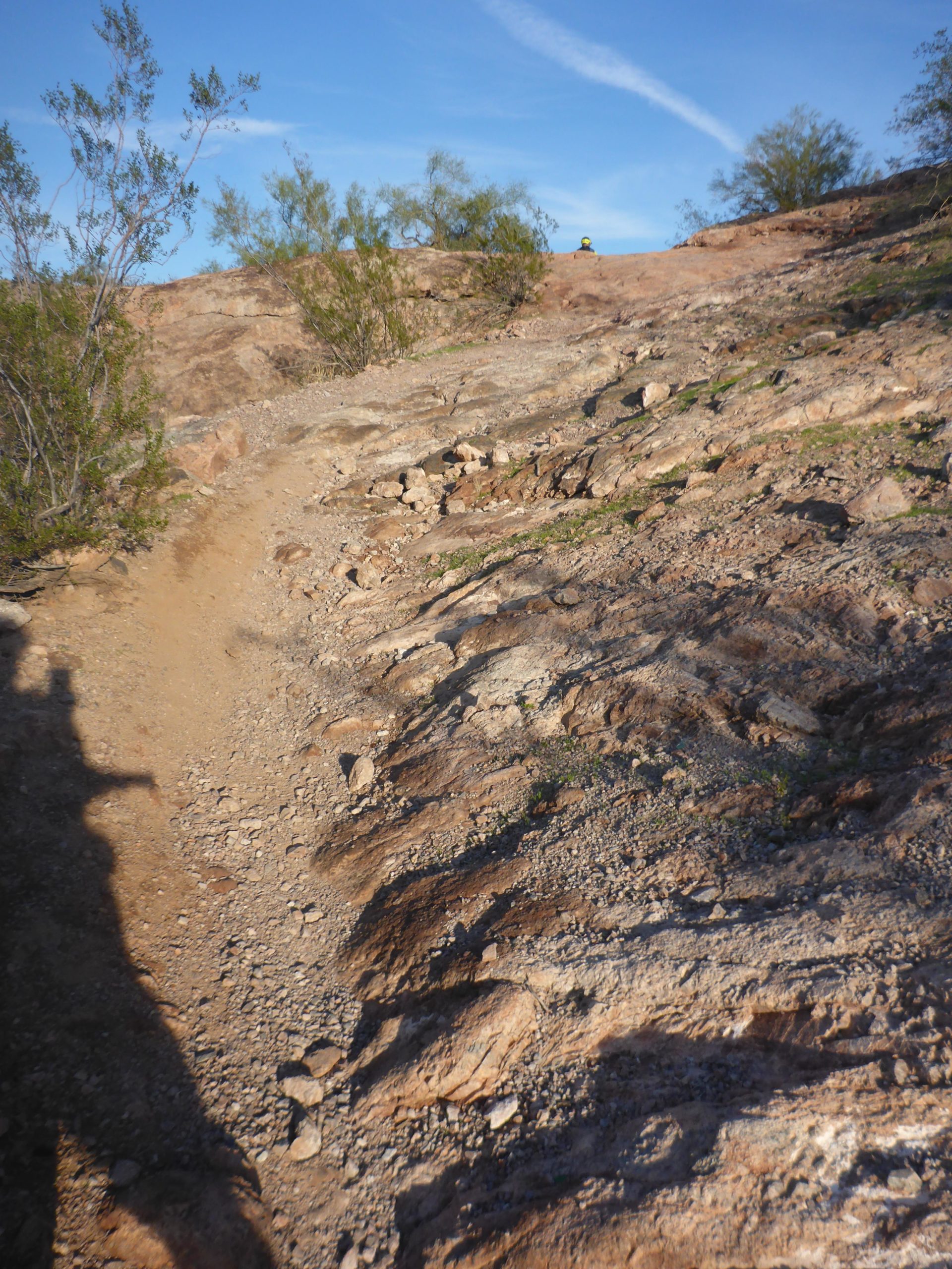 A winding dirt path leads up a rocky hillside, bordered by sparse vegetation. The landscape is bathed in natural light, showcasing the textures of the rocky terrain and a blue sky with scattered clouds above. In the distance, a figure can be seen near the top of the hill, adding a sense of scale to the scene. Papago Park Area mountain bike trail.