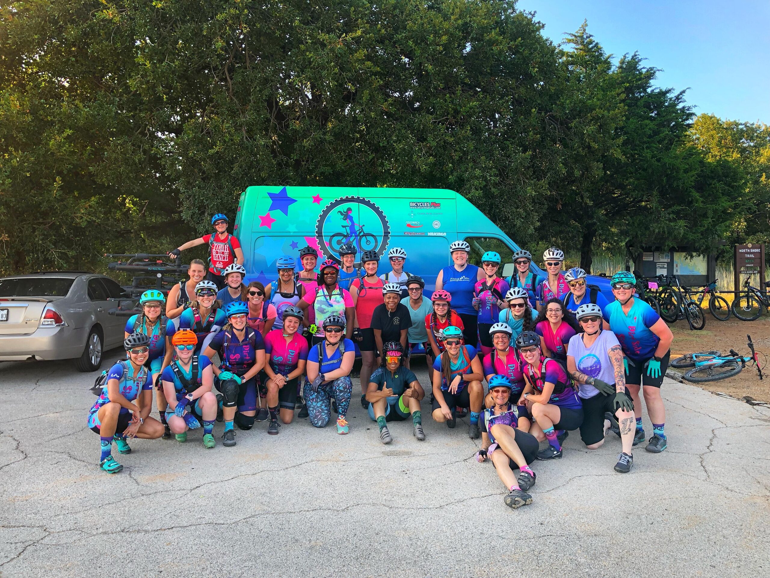 A large group of women posing for a photo in outdoor gear, wearing colorful cycling helmets and jerseys. They are gathered in front of a brightly colored van with a bike-themed logo and parked bicycles nearby. The scene is set in a park-like area with trees in the background, showcasing a vibrant community of cyclists.