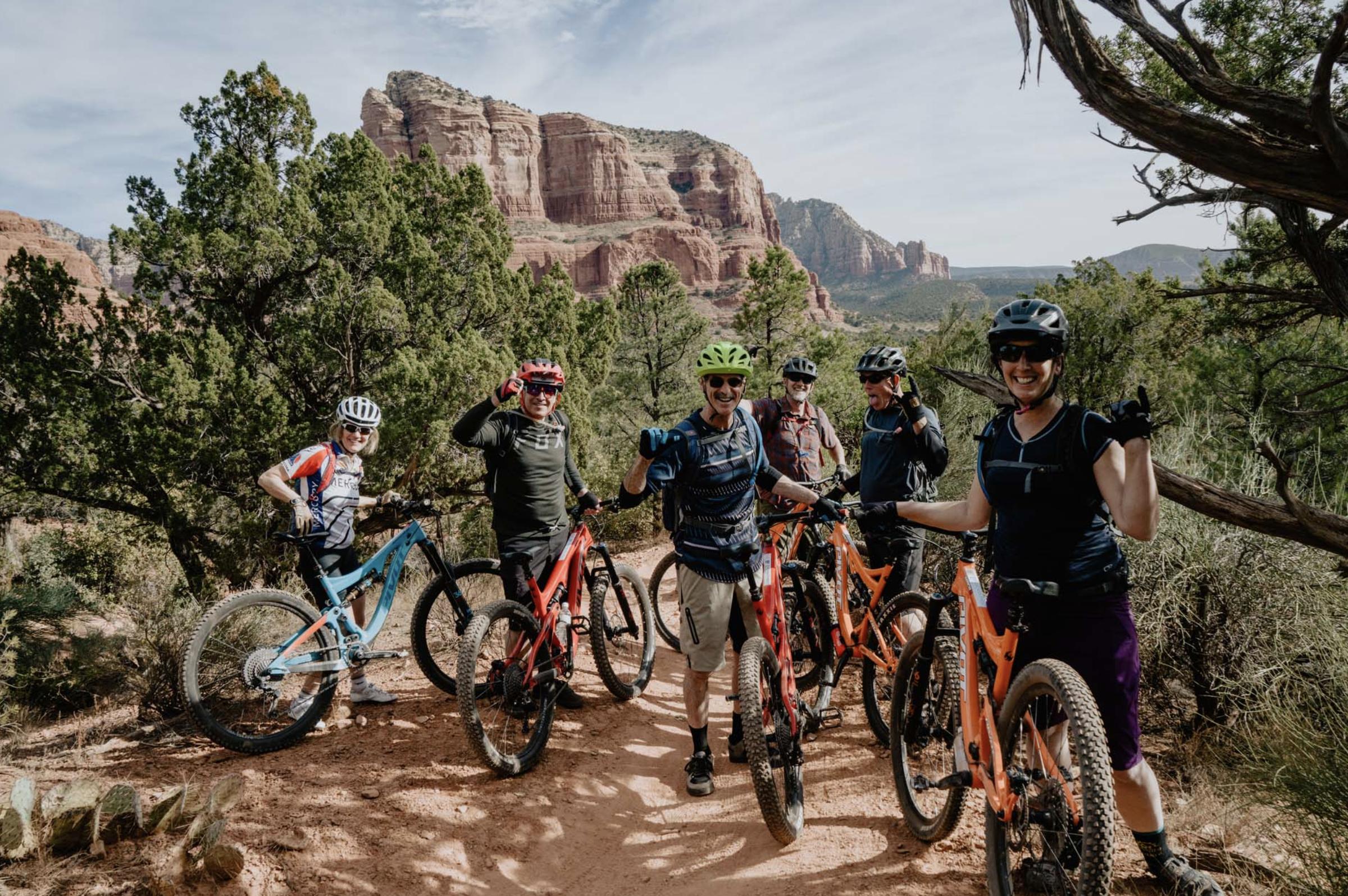A group of six mountain bikers posing together on a dirt trail surrounded by lush greenery and red rock formations in the background. They are wearing helmets and biking gear, with their bicycles parked beside them, and are smiling and making hand gestures to signify enjoyment and camaraderie.