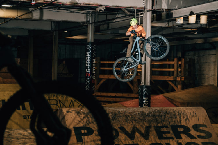 A cyclist performs a mid-air jump on a mountain bike in an indoor bike park. The rider is wearing a bright yellow helmet and an orange shirt. Background features wooden ramps and obstacles with various logos and markings on them, illuminated by soft indoor lighting.
