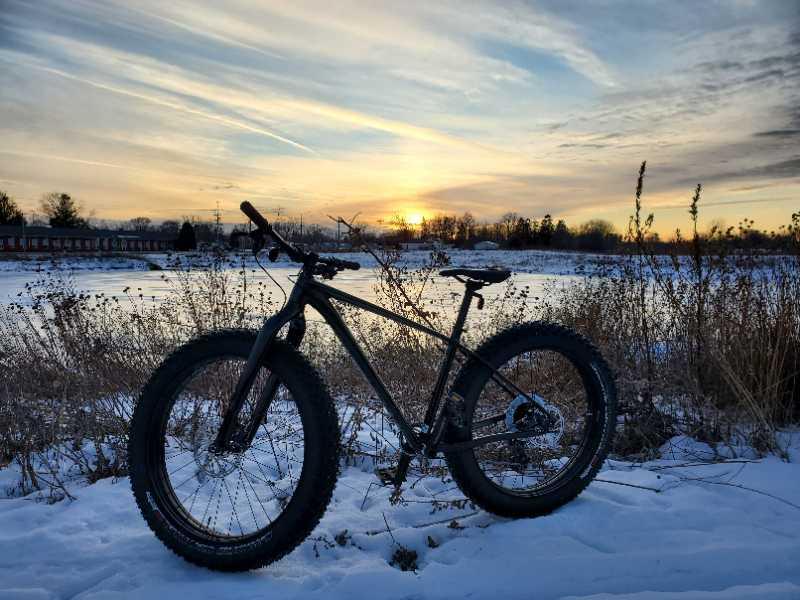 A black fat bike stands on a snowy landscape, with a frozen body of water and a sunset in the background. The sky is filled with soft clouds, showcasing warm hues of orange and yellow as the sun sets behind the horizon. Tall grass and plants surround the bike, adding a natural touch to the serene winter scene. Appleton Memorial Park mountain bike trail.