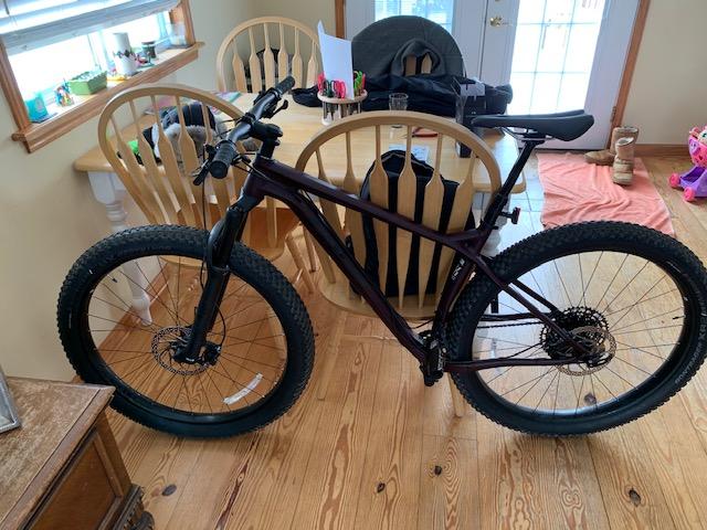 Trek Stache 7: A mountain bike with thick tires is leaning against a wooden dining table in a well-lit room. In the background, there are several wooden chairs and a door leading outside. The floor is made of wooden planks, and there are personal items visible, including a backpack and a pair of boots.