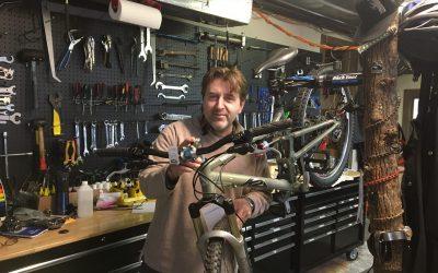 A person in a workshop holding a bicycle, with various tools and bike maintenance equipment visible on the wall and workbench behind them.