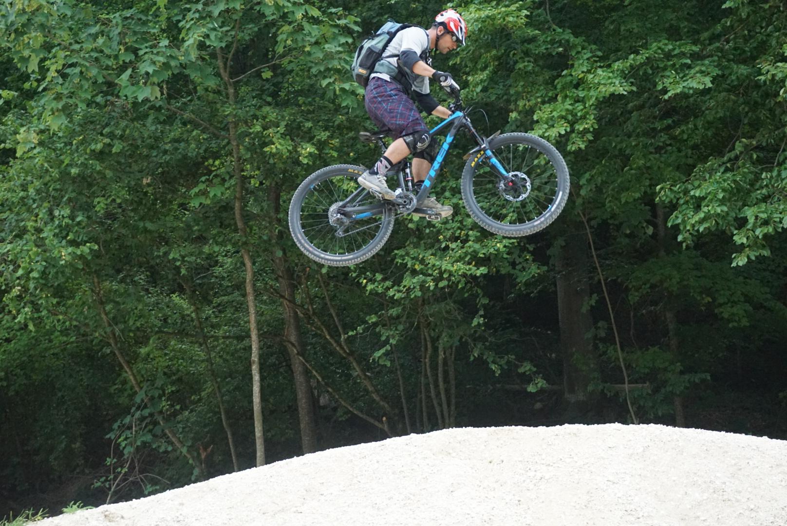 A mountain biker mid-air while performing a jump over a sandy ramp, surrounded by lush green trees. The cyclist is wearing a helmet and protective gear, showcasing agility and skill in a natural outdoor setting. SWU Trails mountain bike trail.