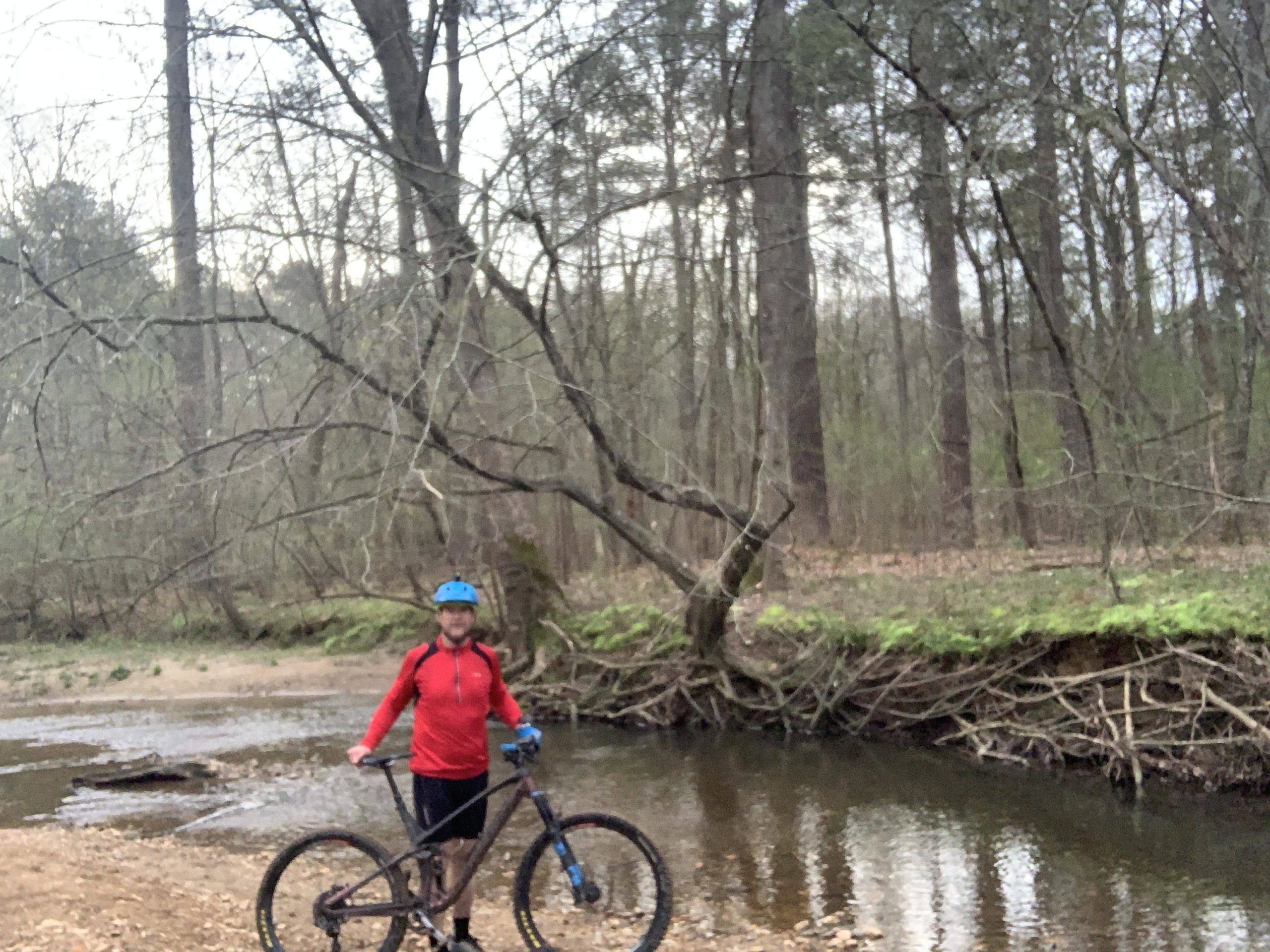A person in a red long-sleeve shirt and blue helmet stands beside a mountain bike near a small creek in a wooded area. The background features tall trees with bare branches and green foliage along the water's edge. The scene is calm and natural, suggesting a recreational outing. Blankets Creek mountain bike trail.