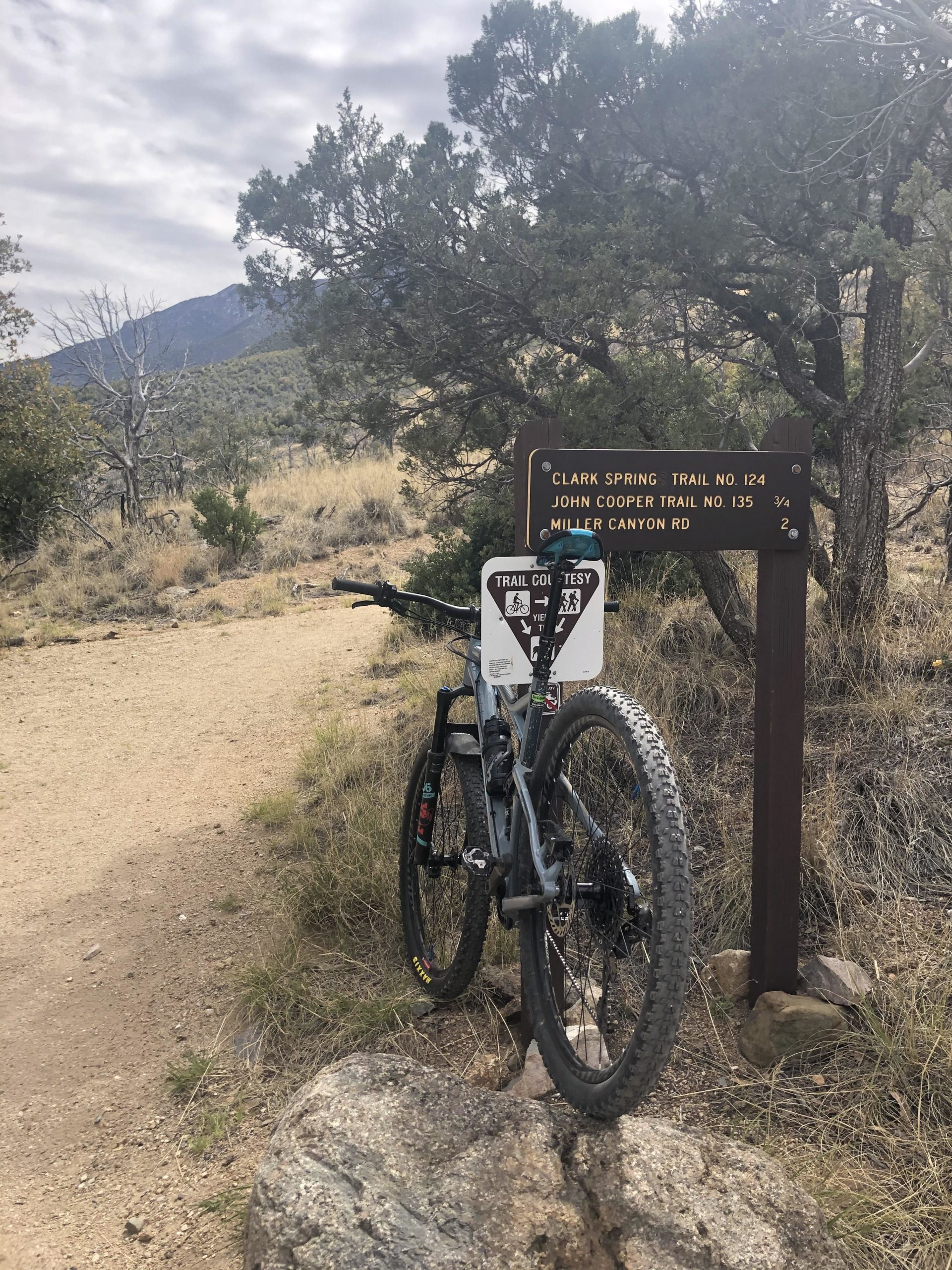 A mountain bike leaning against a trail sign indicating directions for Clark Spring Trail No. 124, John Cooper Trail No. 135, and Miller Canyon Road. The background features a rocky terrain with sparse vegetation and mountains under a cloudy sky. Perimeter Trail mountain bike trail.