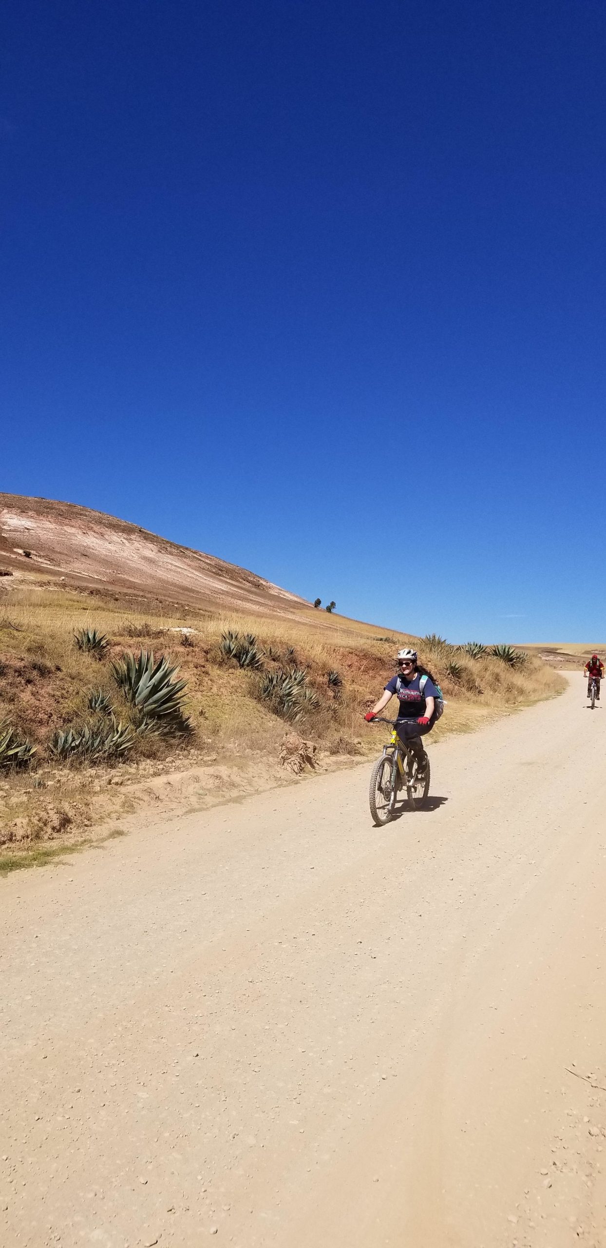 A cyclist riding on a dirt path with a clear blue sky overhead. The landscape includes rolling hills and sparse vegetation along the sides of the path. Another cyclist is visible in the background. Sacred Valley Downhill mountain bike trail.