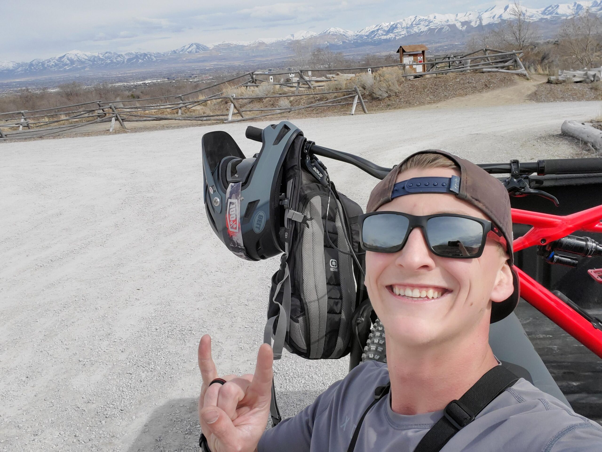 Specialized Enduro Comp 29: A smiling person wearing sunglasses and a cap, holding up a hand gesture while standing next to a mountain bike in a scenic outdoor location. In the background, snow-capped mountains and a wooden fence are visible, with a cloudy sky above. A backpack and a helmet are also visible near the bike.