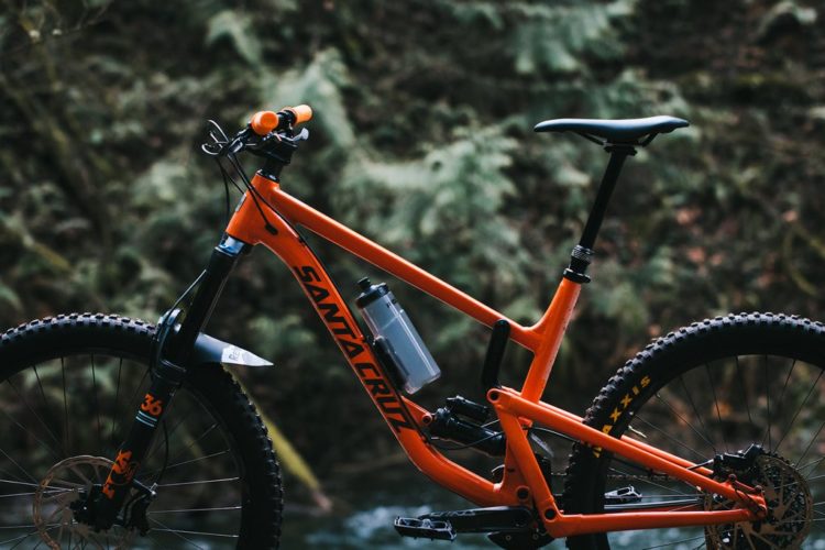Alt text: A close-up view of an orange Santa Cruz mountain bike positioned next to a river, surrounded by lush greenery. The bike features a large front tire, a water bottle attached to the frame, and a sleek design with visible suspension components.