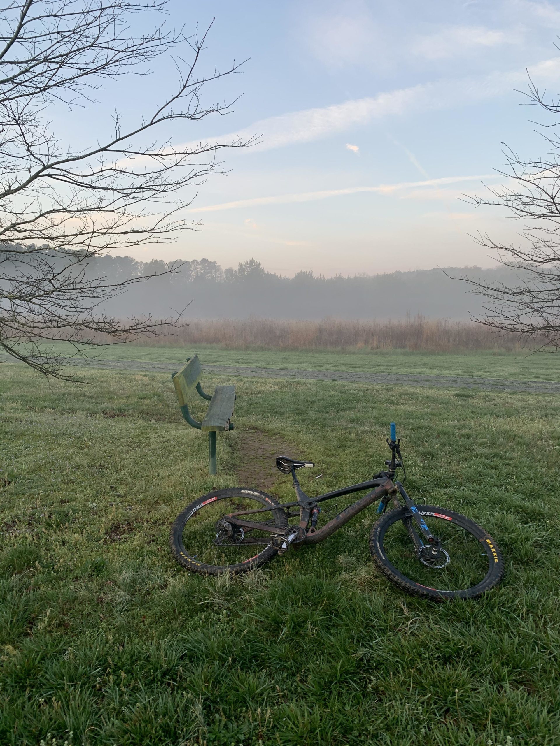 A mountain bike rests on the grass near a green park bench, with a misty landscape and a serene sky in the background. The scene is calm, with bare trees framing the view, and a fog settling over the distant fields. Allatoona Creek Park mountain bike trail.