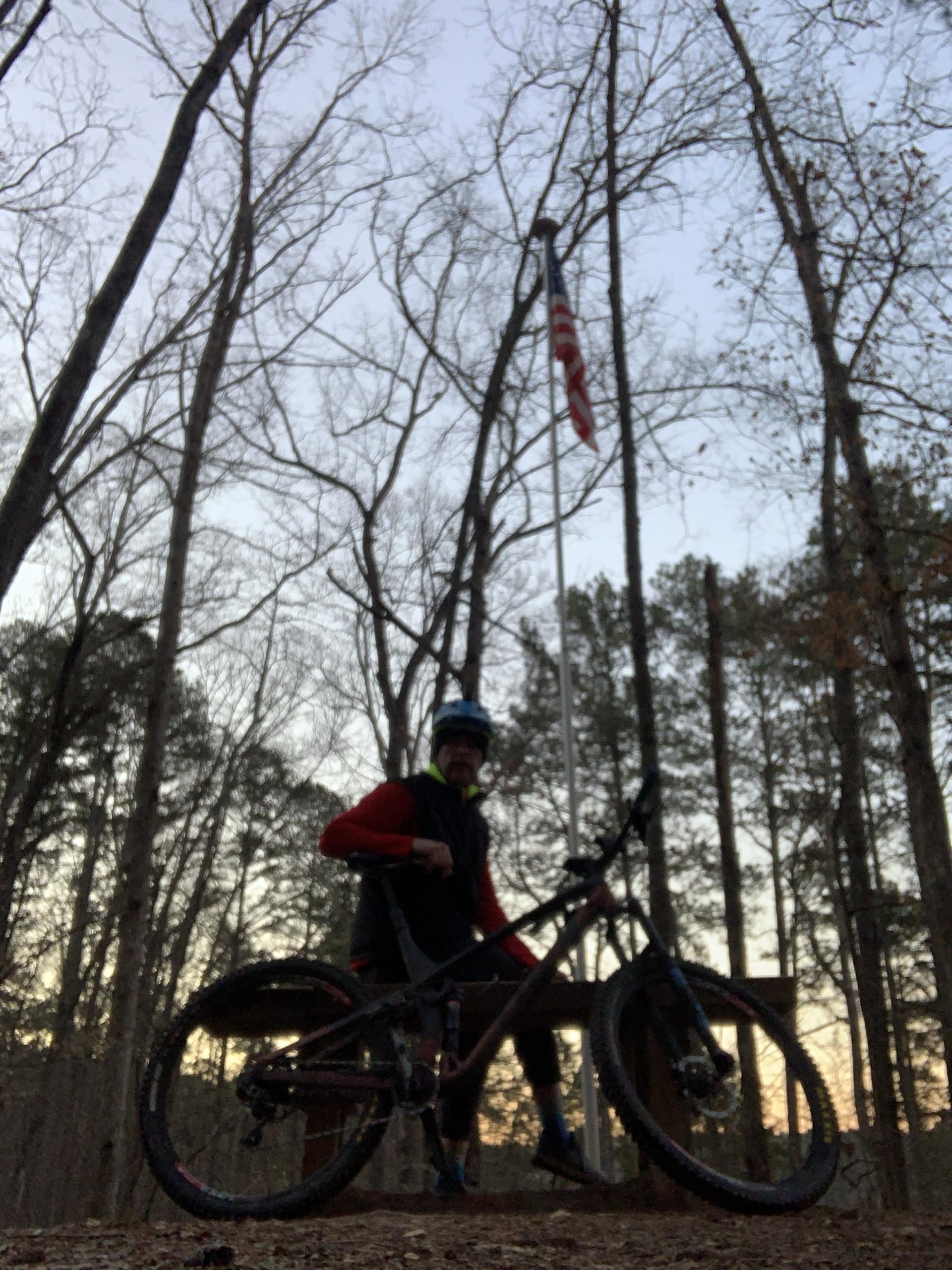 A cyclist sitting on their mountain bike in a forested area during dusk, with bare trees in the background and an American flag visible on a nearby flagpole. The cyclist is wearing a helmet and a red jacket, and the scene captures a peaceful outdoor ambiance. Blankets Creek mountain bike trail.