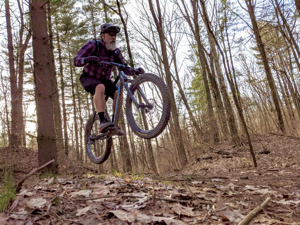 A man with a beard wearing a helmet and plaid shirt performs a jump on a mountain bike while riding through a wooded area with trees in the background and leaves on the ground. Alpha mountain bike trail.