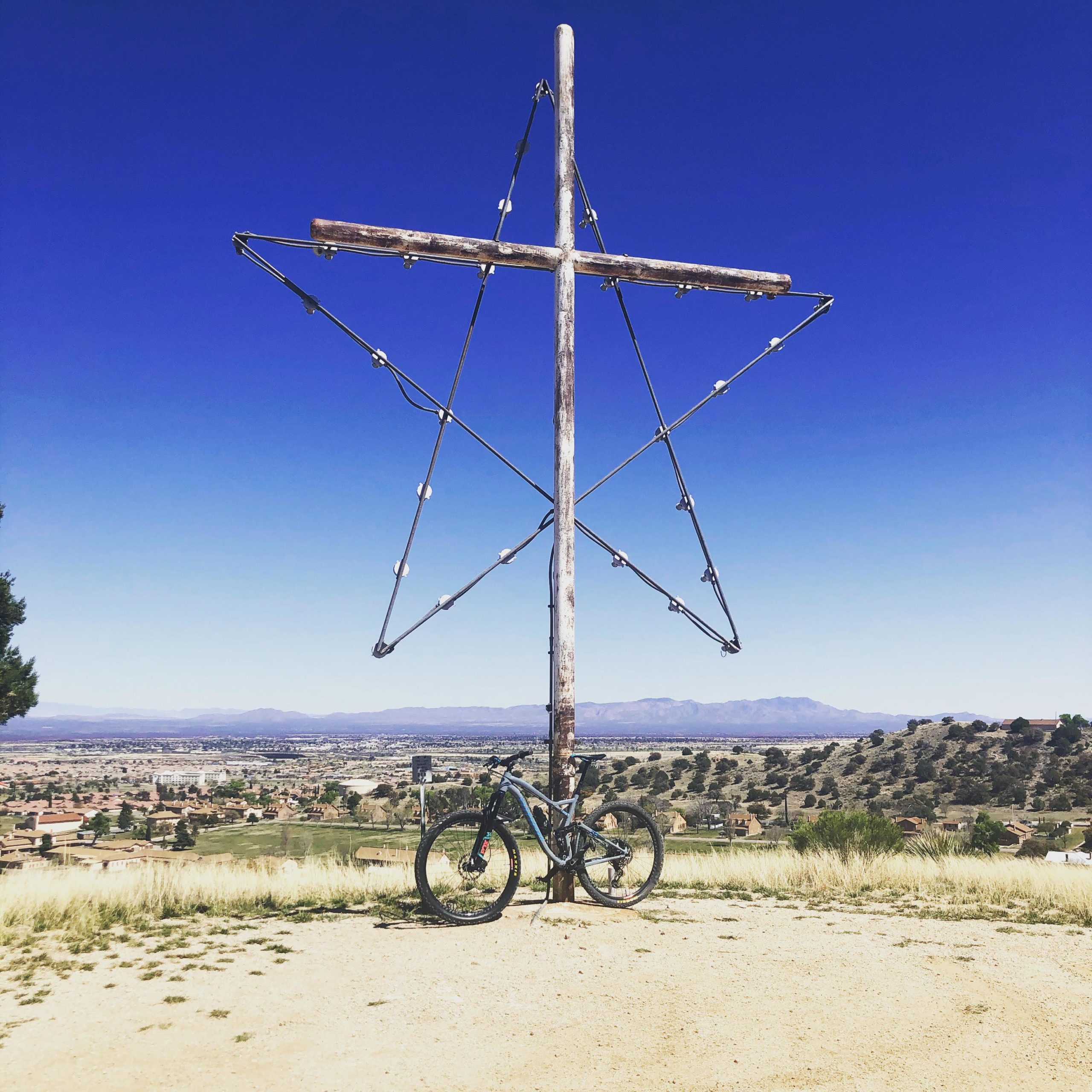 A wooden star structure standing on a hill, with a mountain bike leaning against its base. The backdrop features a clear blue sky and a view of a valley with houses and greenery below. Wren Arena Red Loop Trail mountain bike trail.