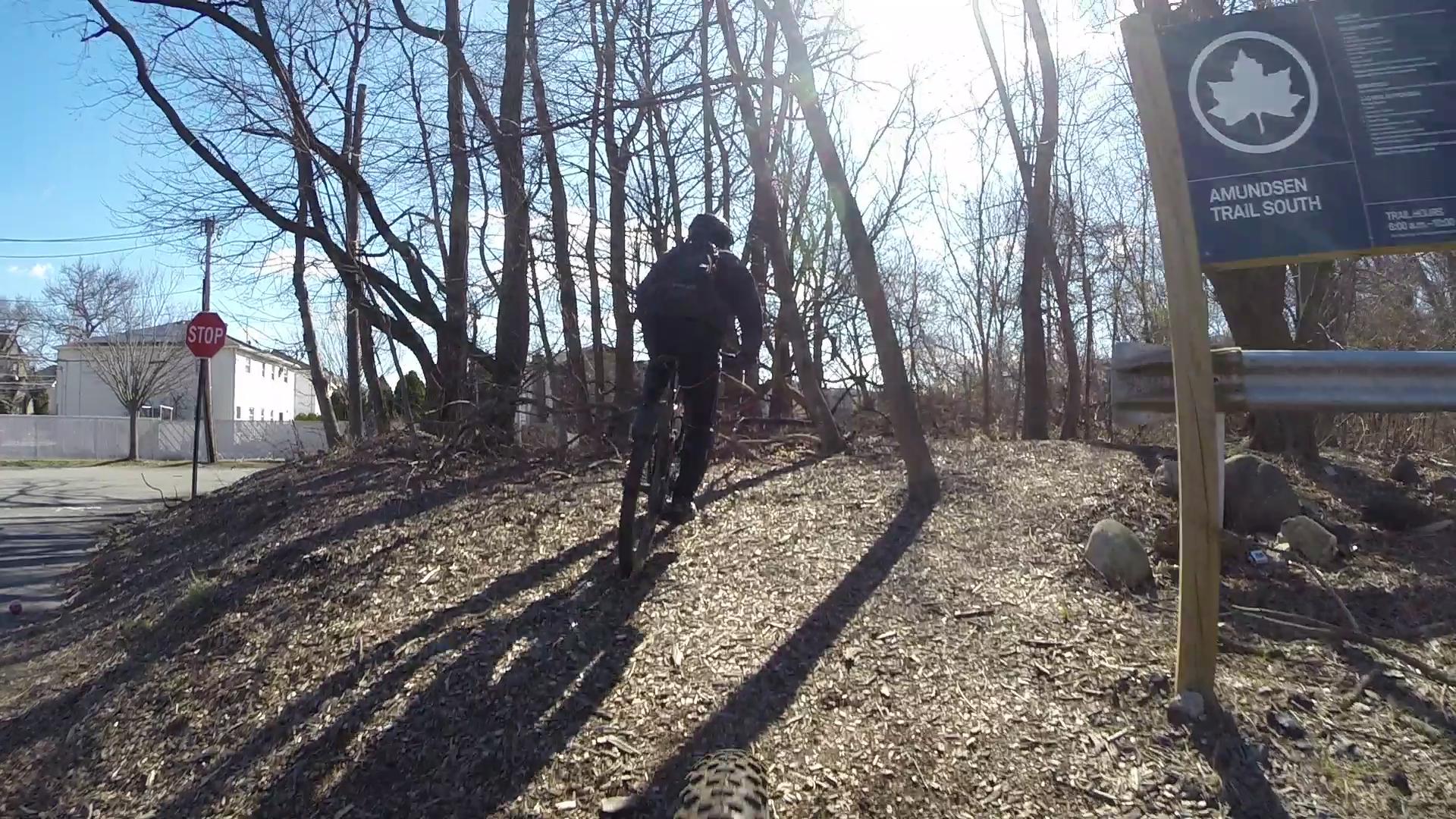 A mountain biker riding up a dirt trail surrounded by bare trees on a sunny day, with a "STOP" sign and an "Amundsen Trail South" sign visible in the background. Richmond Avenue and Forest Hill road mountain bike trail.