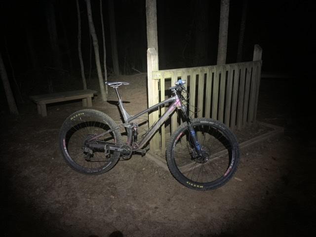 A mountain bike with fat tires is parked near a wooden bench and a bike rack in a wooded area at night. The scene is illuminated by a bright light, highlighting the bike's features and the surrounding trees. Blankets Creek mountain bike trail.