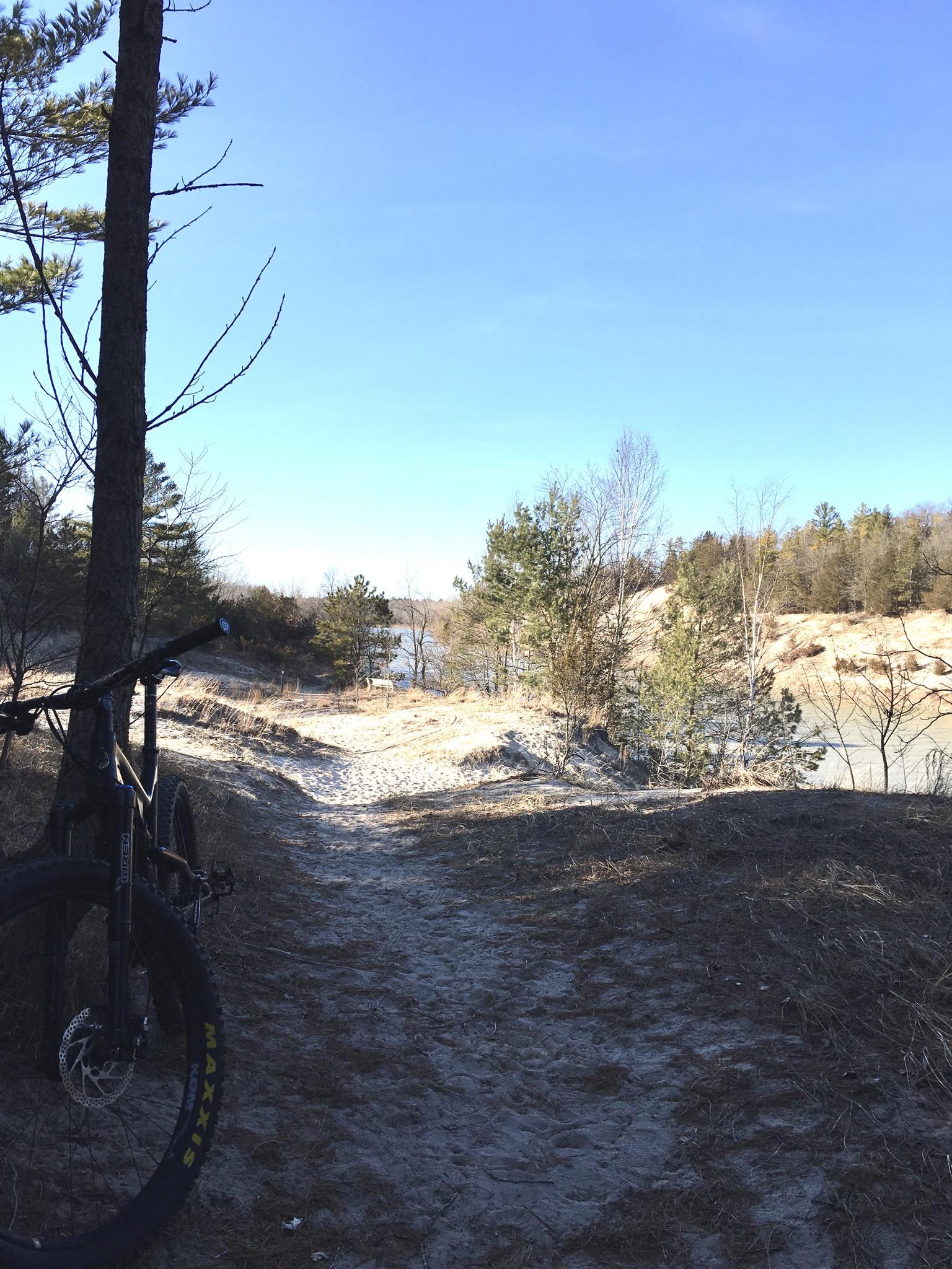 A winding sandy path along a river, bordered by trees and shrubs, with a mountain bike leaning against a tree in the foreground. The sky is clear and blue, indicating a sunny day. Lambton County Heritage Forest mountain bike trail.